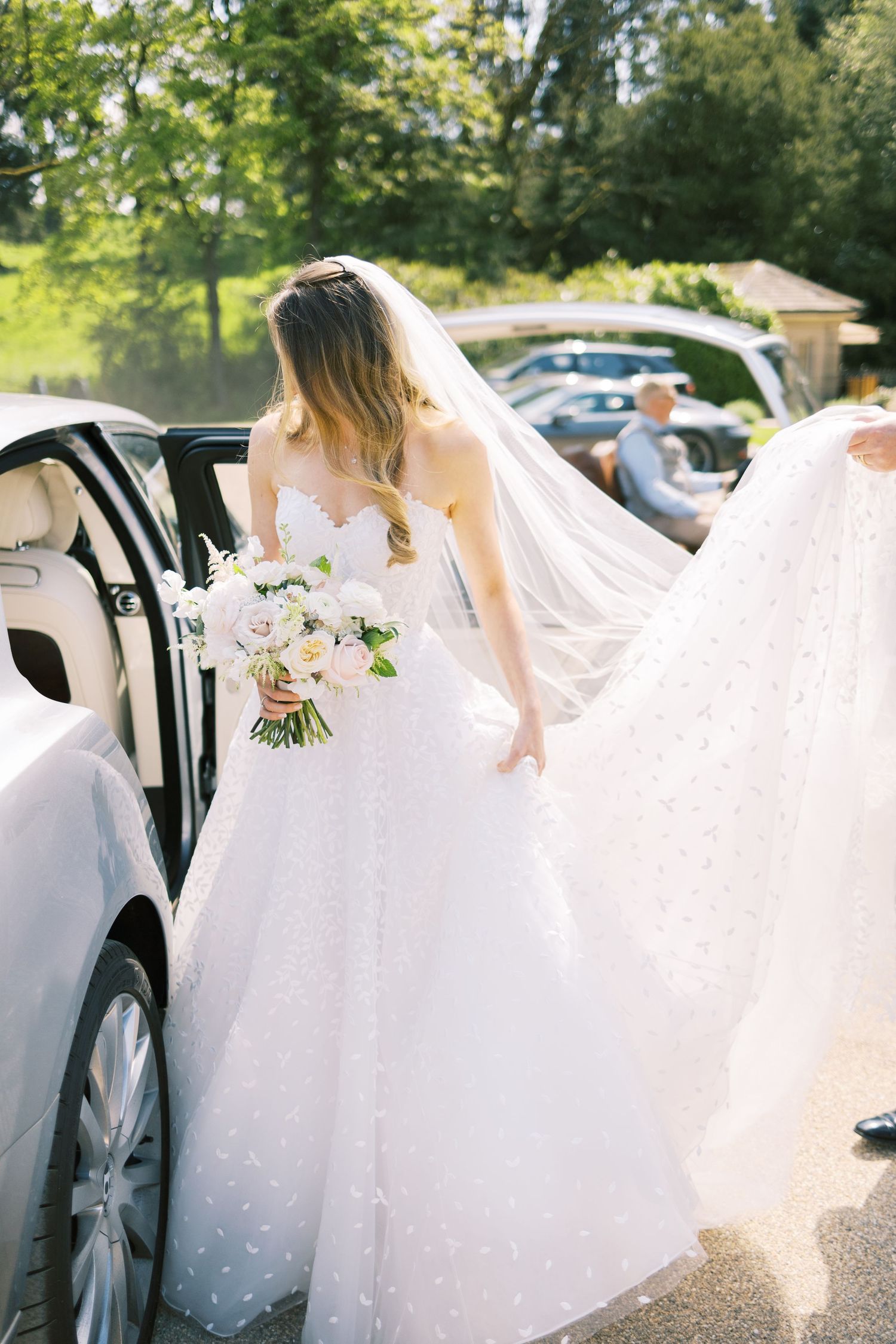 Bride in polka dot ballgown with veil flowing beside white car.