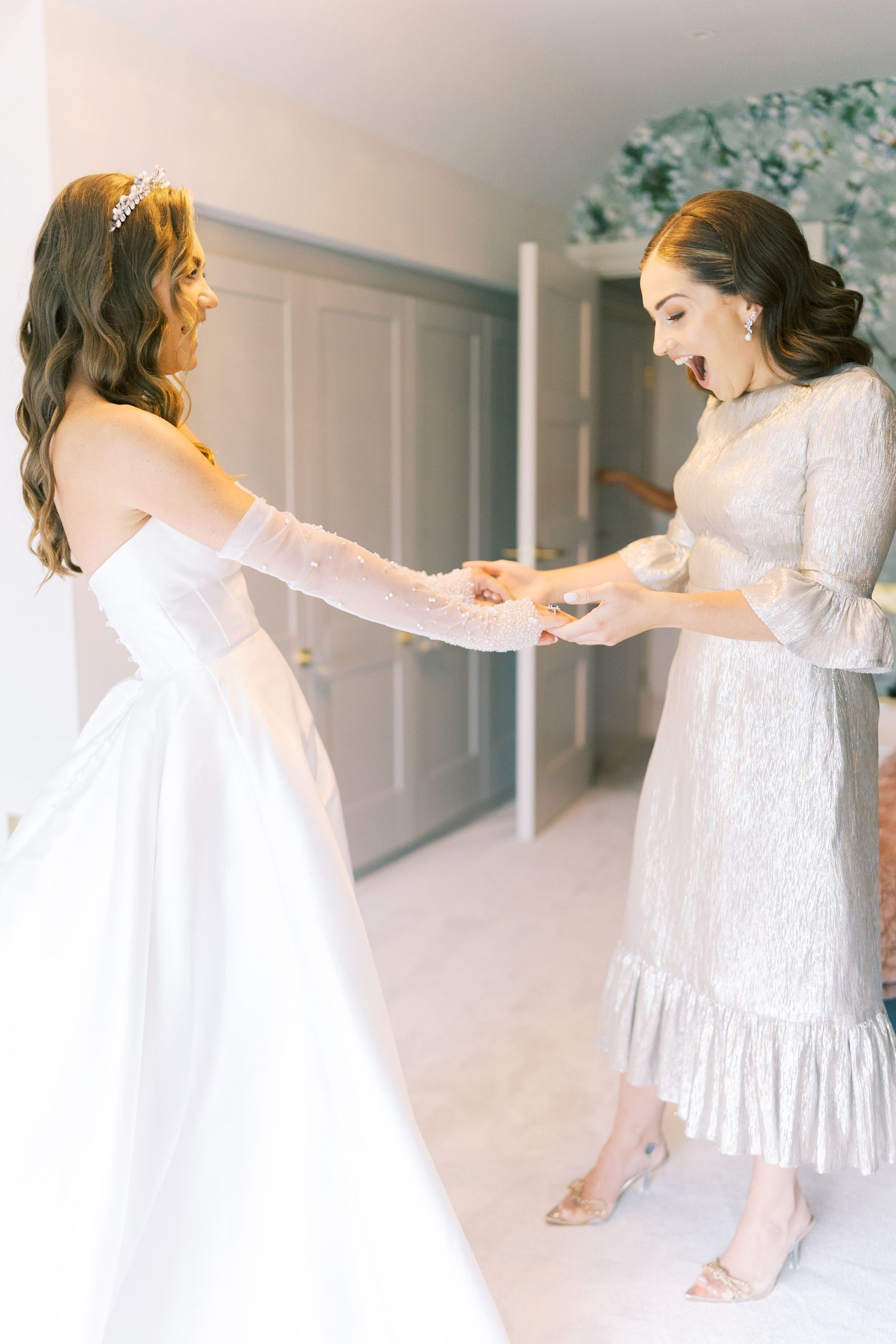 Bride in white wedding gown excitedly holds hands with bridesmaid in silver lace dress during bridal preparations.