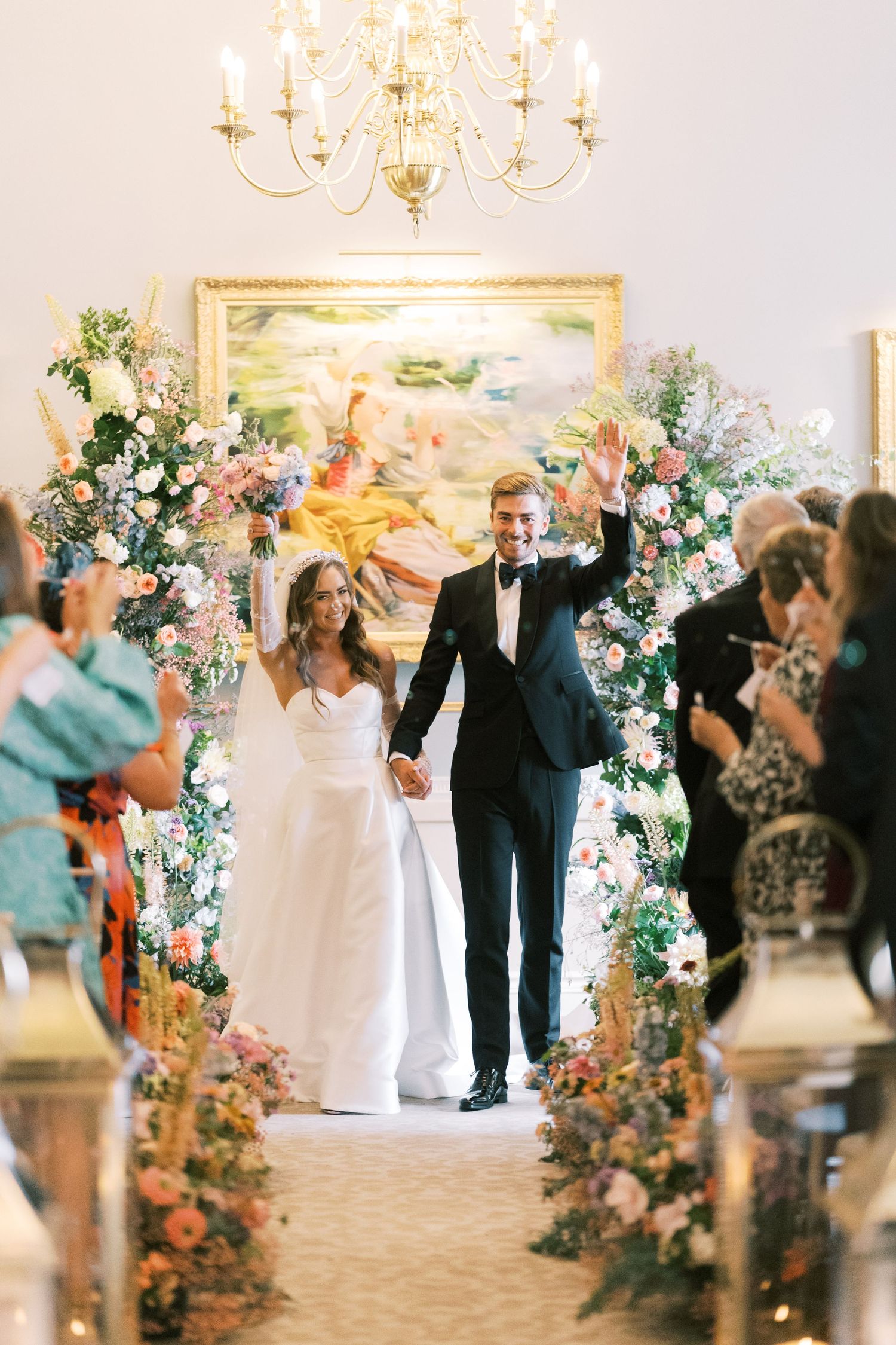 Bride and groom wave joyfully while walking down floral-lined aisle after wedding ceremony indoors.