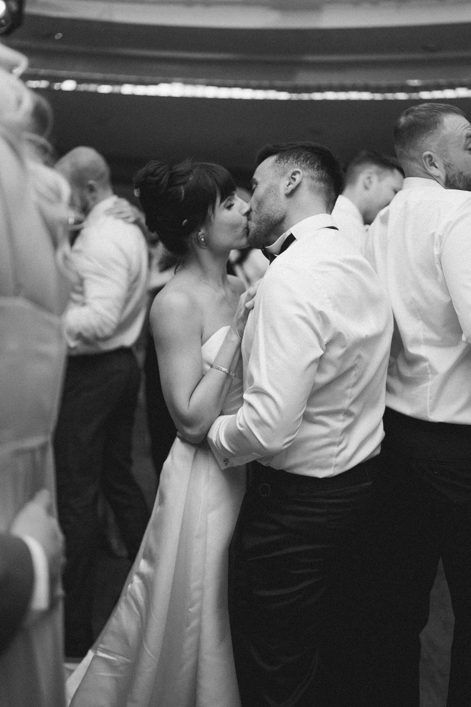 Black and white photo of bride and groom sharing a kiss on the dance floor surrounded by wedding guests.