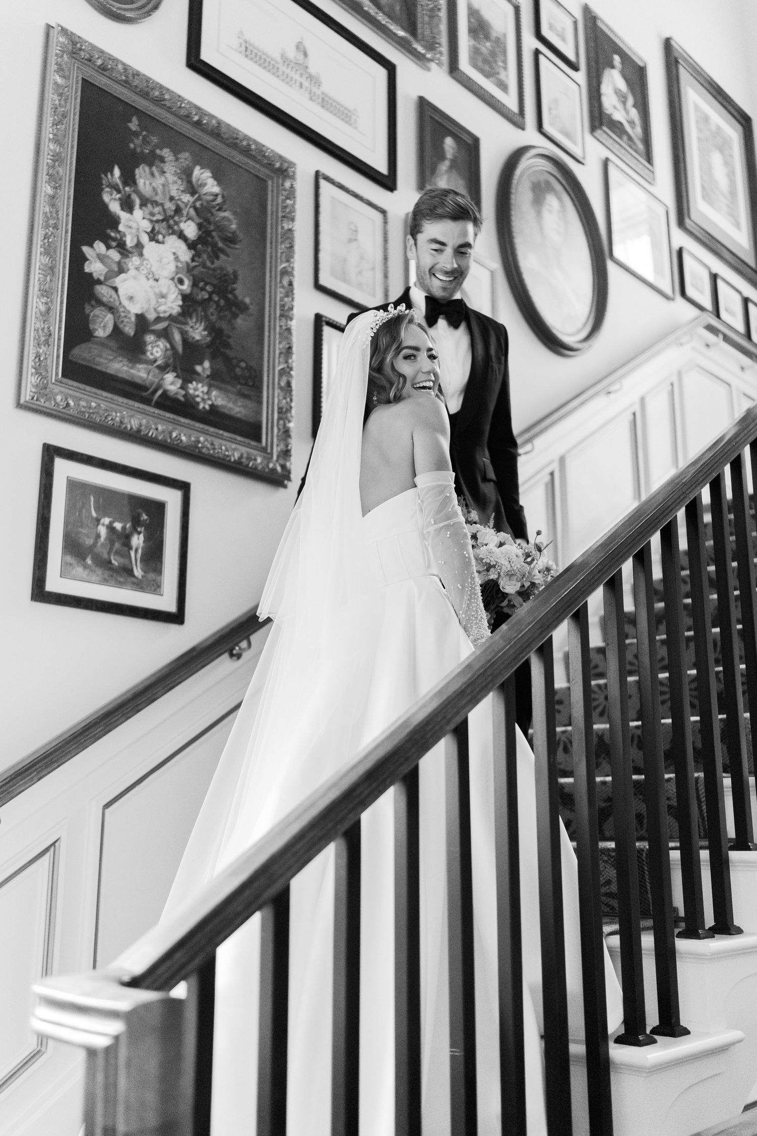 Black and white photo of a smiling bride and groom on an elegant staircase adorned with framed artwork on the wall.