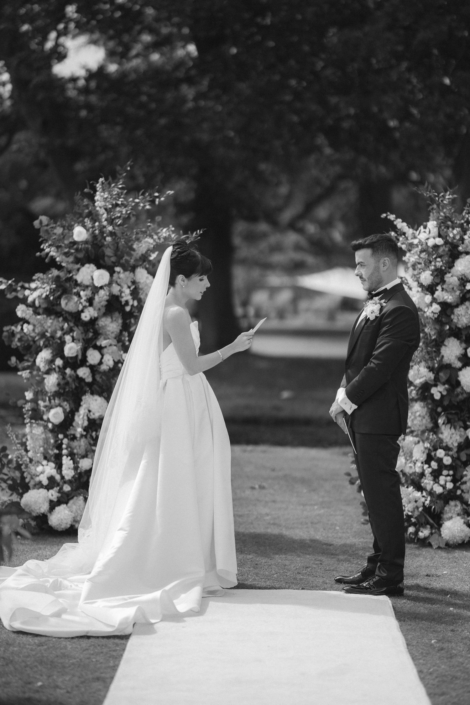 Bride reads wedding vows to groom at outdoor ceremony flanked by floral arrangements, black and white photo.