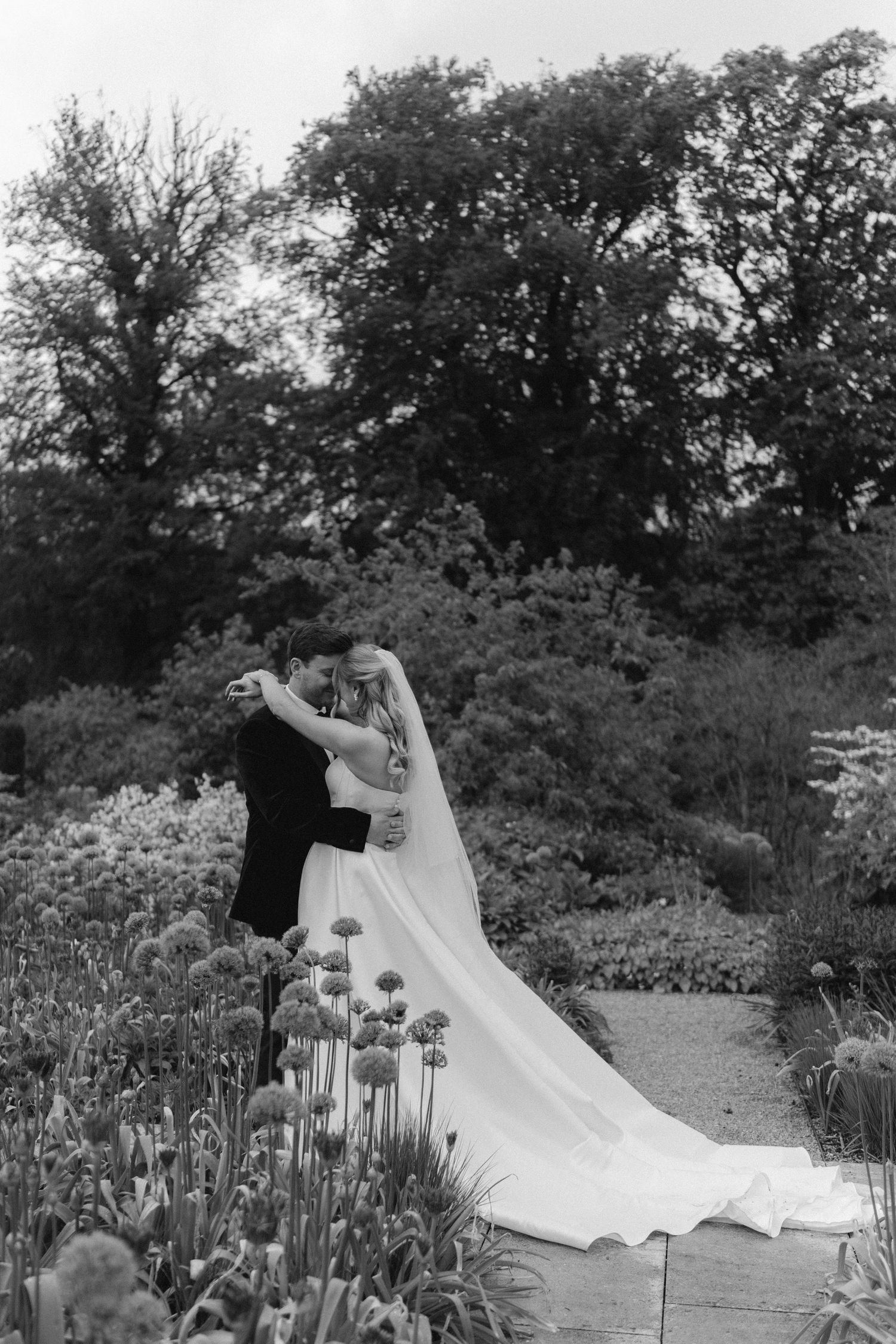 Black and white photo of bride and groom embracing in a lush garden, wedding veil trailing among tall flowers.