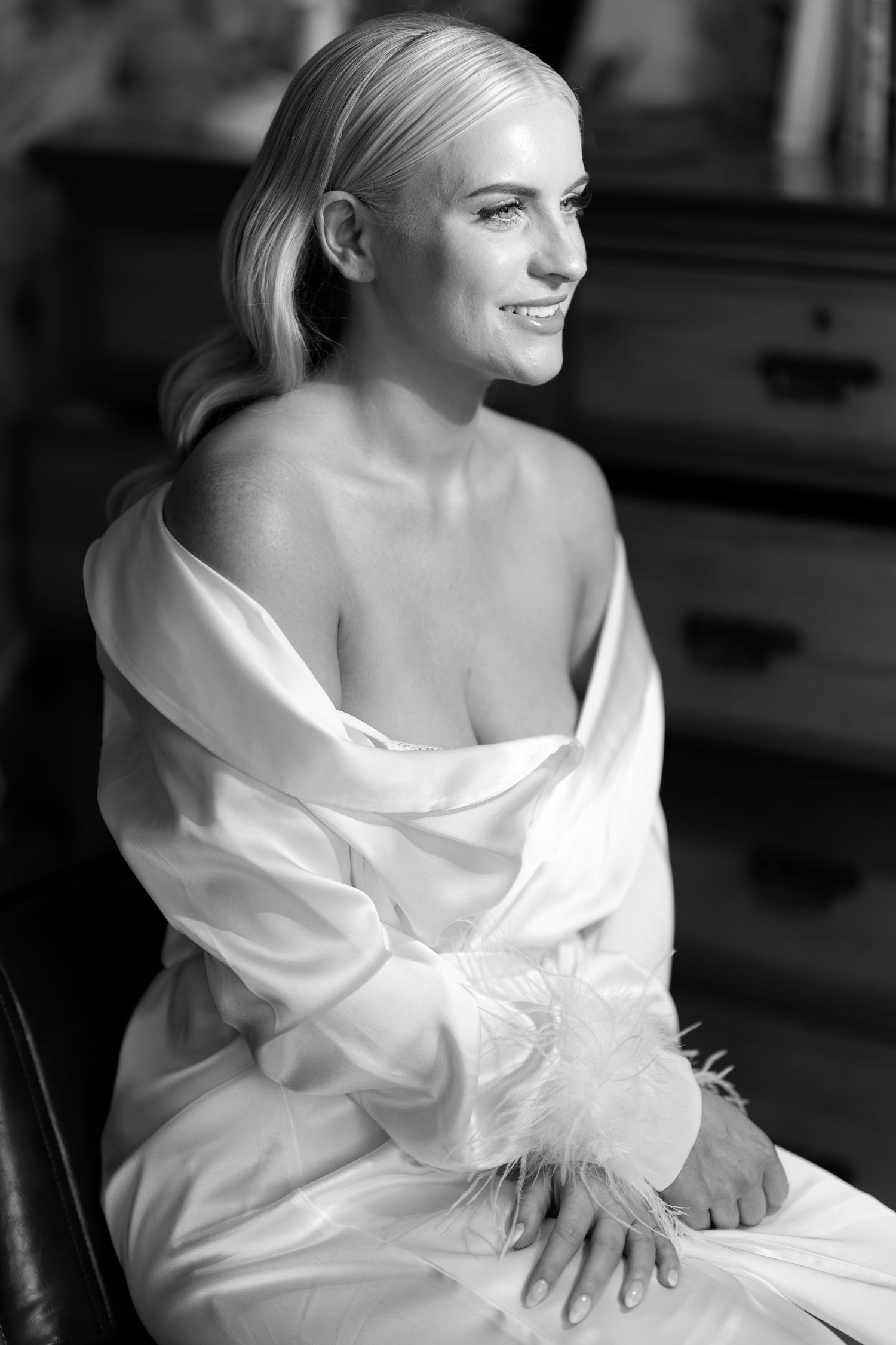 Black and white portrait of a smiling blonde bride in a white silk robe with feather cuffs.