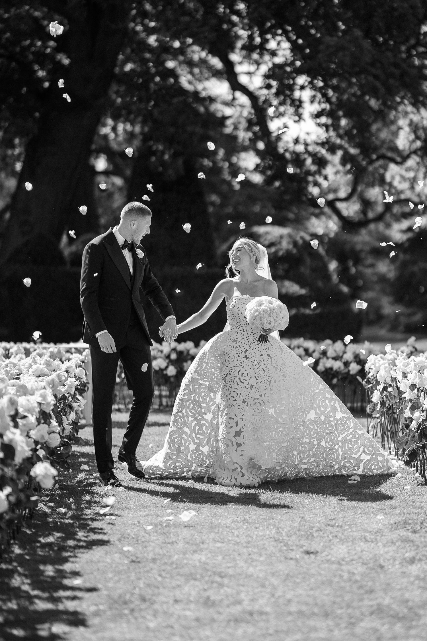 Black and white photo of bride and groom walking through a flower-lined aisle with petals falling.