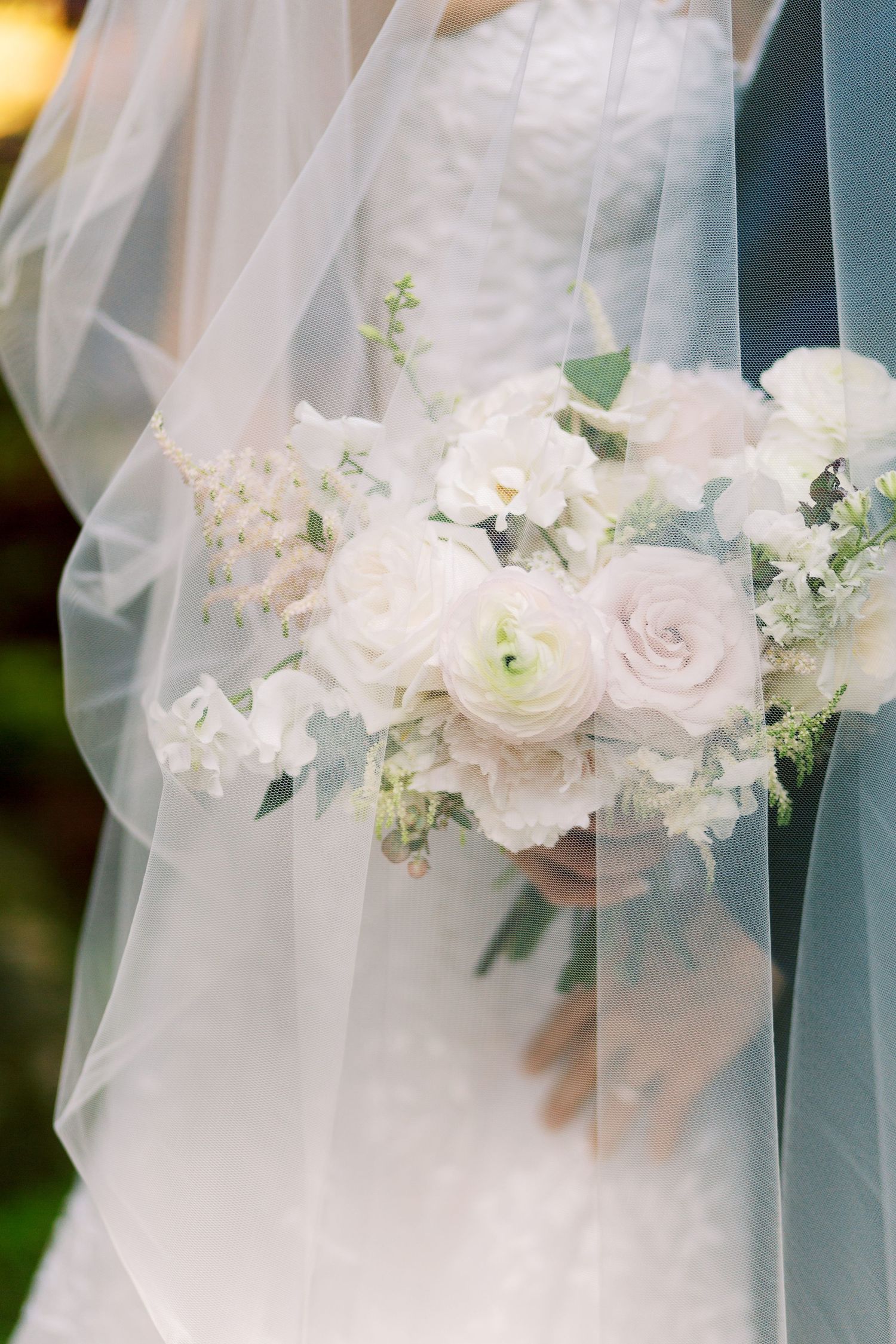 Bride holding a lush white and blush rose bouquet with greenery, draped in a flowing sheer wedding veil.