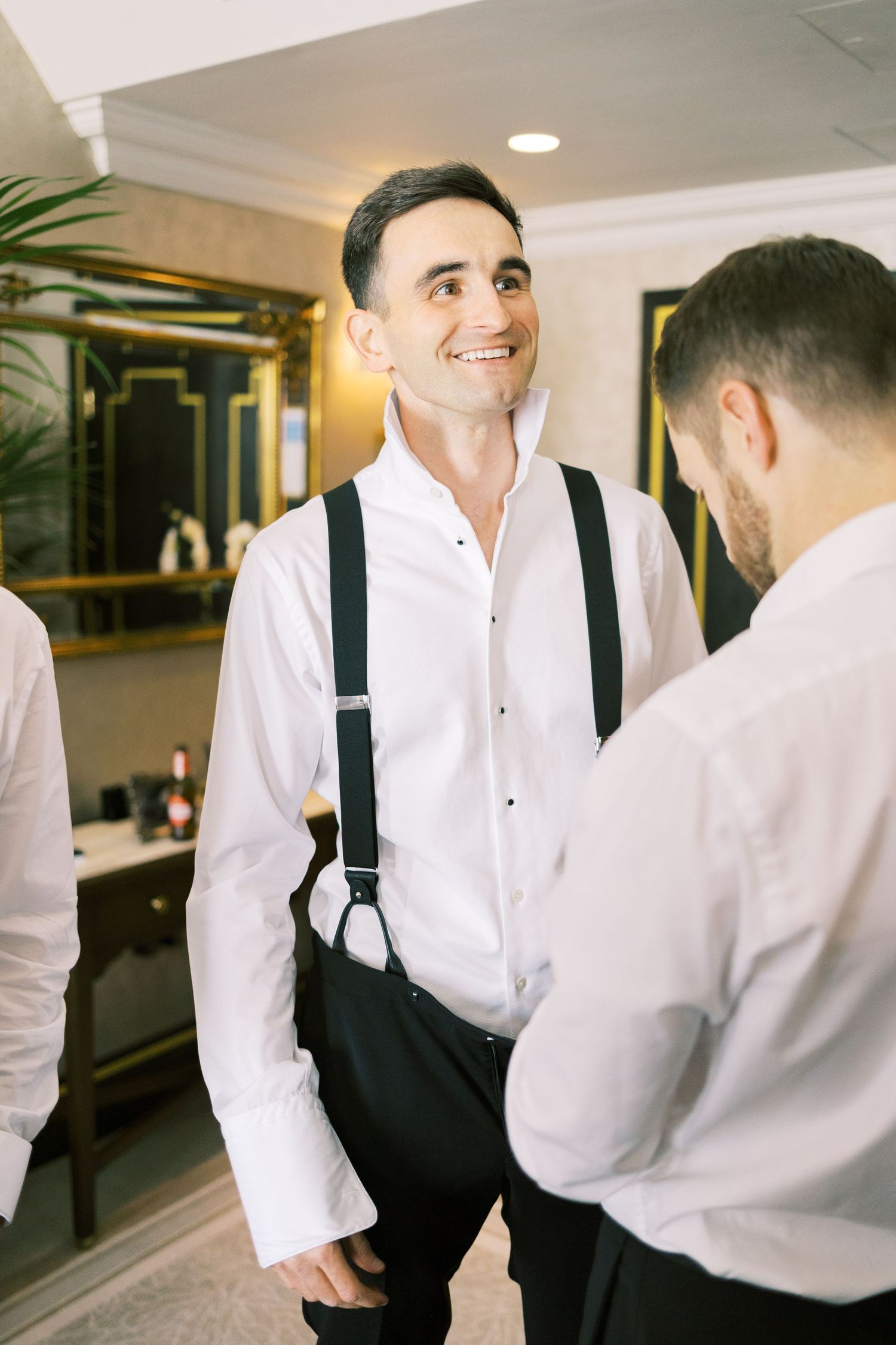 Smiling groom in white shirt and suspenders chatting with groomsman in stylish hotel room.