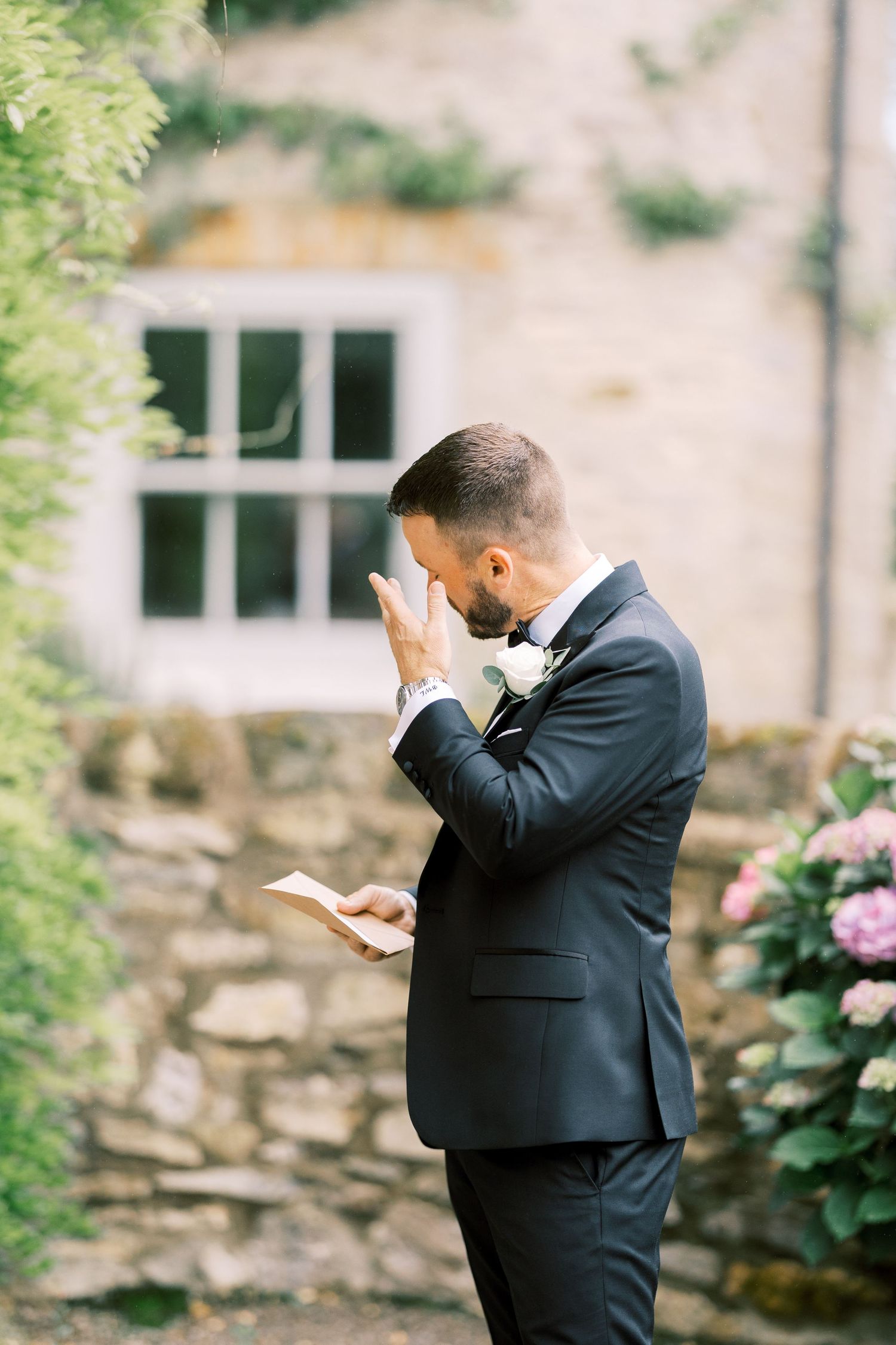 Emotional groom in black suit reads wedding vows, wiping tears outside stone venue with pink flowers.