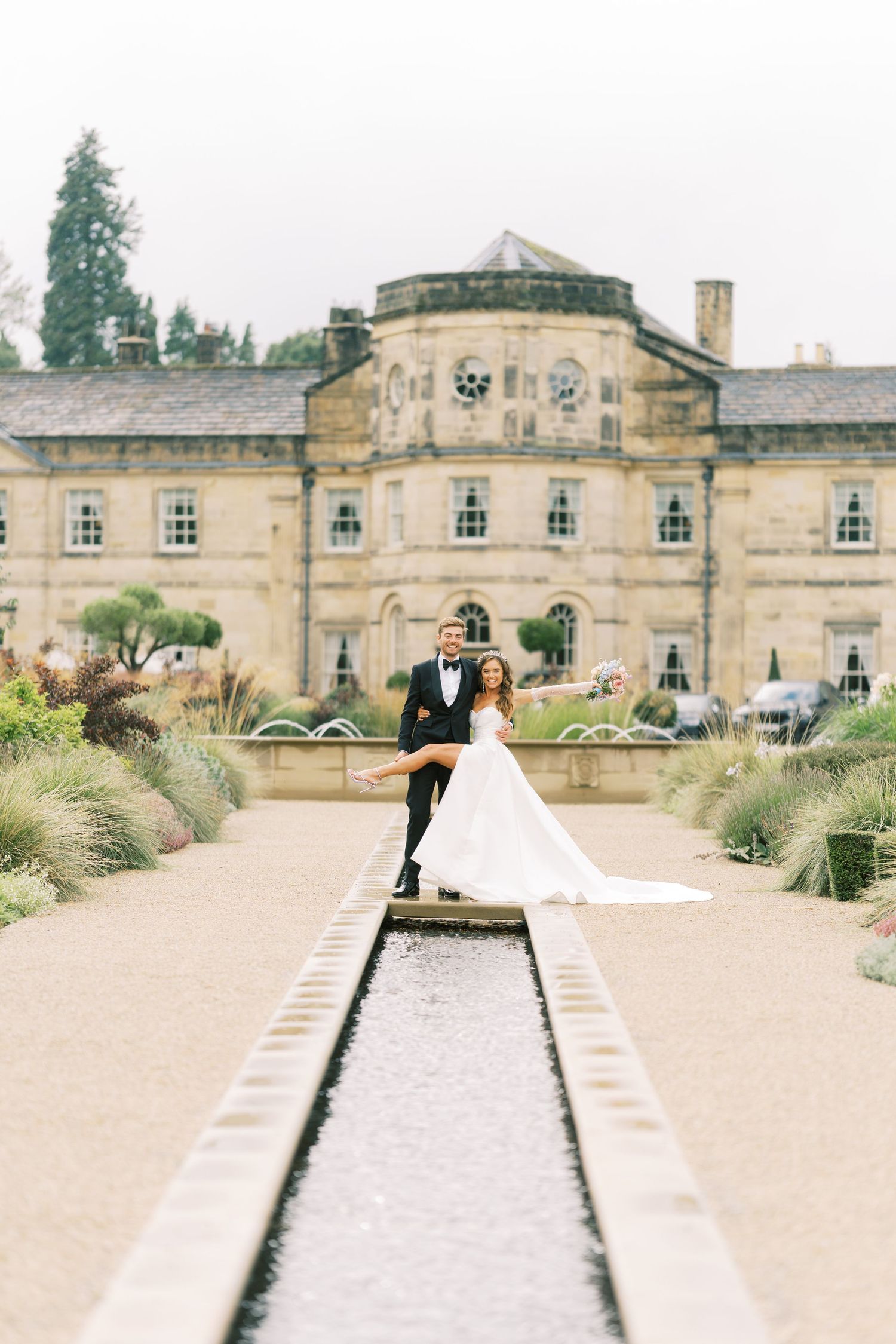 Bride and groom pose elegantly in front of grand stone manor beside a long reflecting pool in manicured gardens.