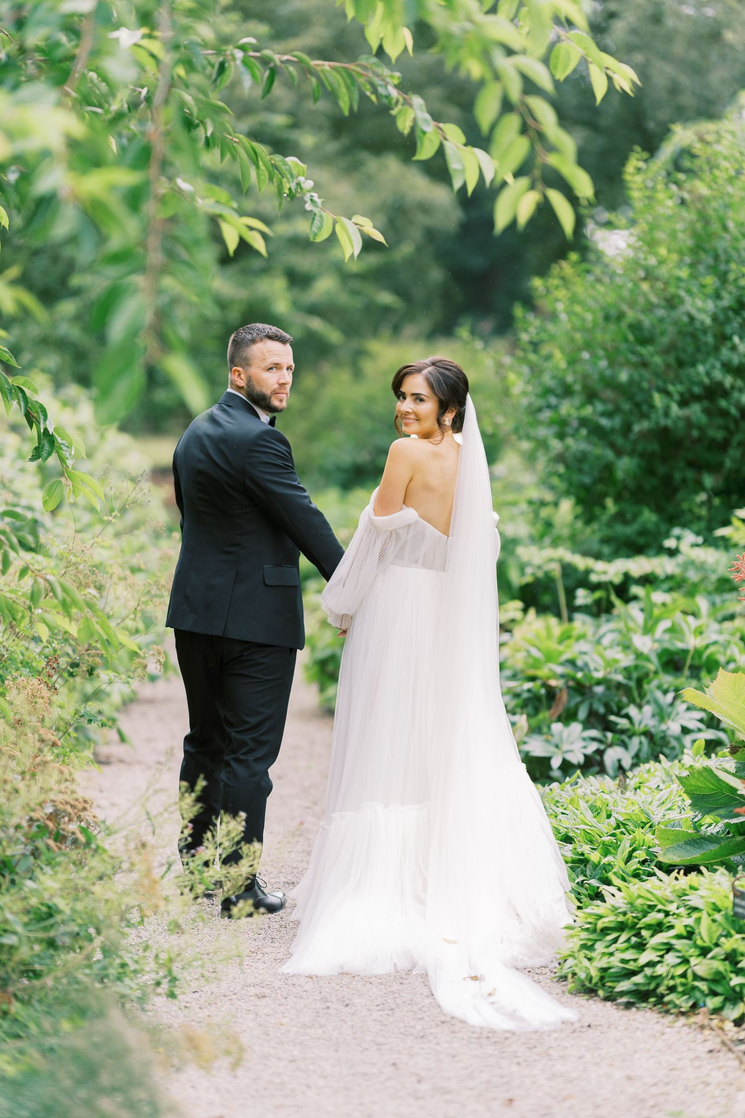 A bride and groom hold hands on a garden path, she in a white gown with veil, he in a black suit.