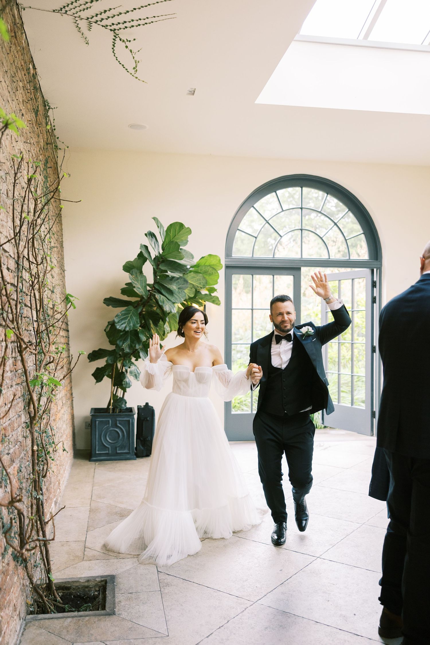 Bride and groom wave joyfully as they enter their wedding reception through an elegant sunlit venue.