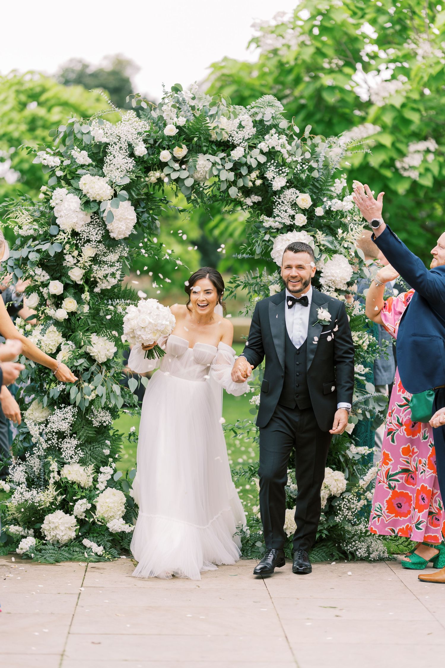 Bride and groom exit ceremony under white floral arch, holding hands and smiling as guests celebrate with confetti.