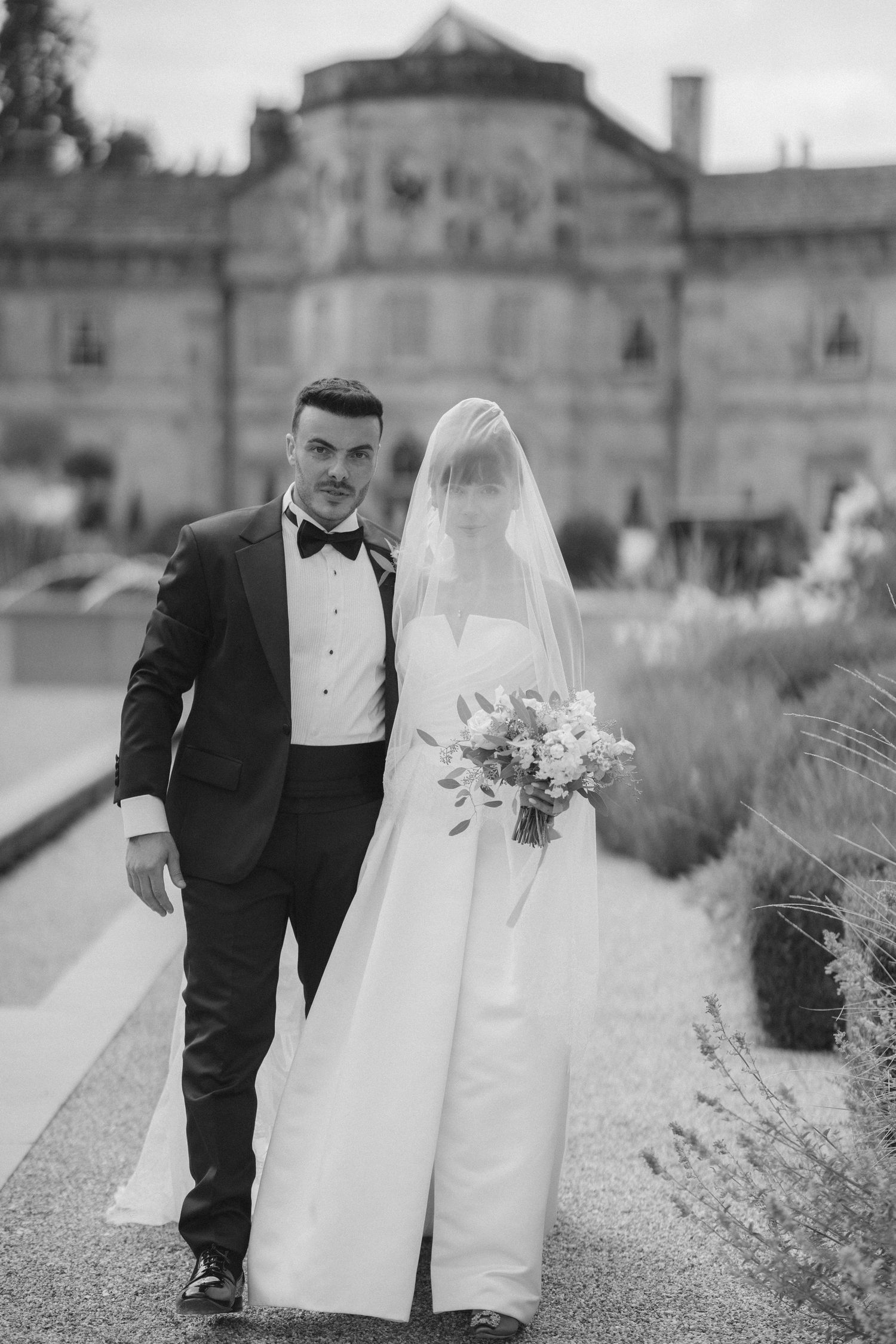 Black and white photo of a bride and groom walking outdoors near a grand building, bride holding a bouquet.