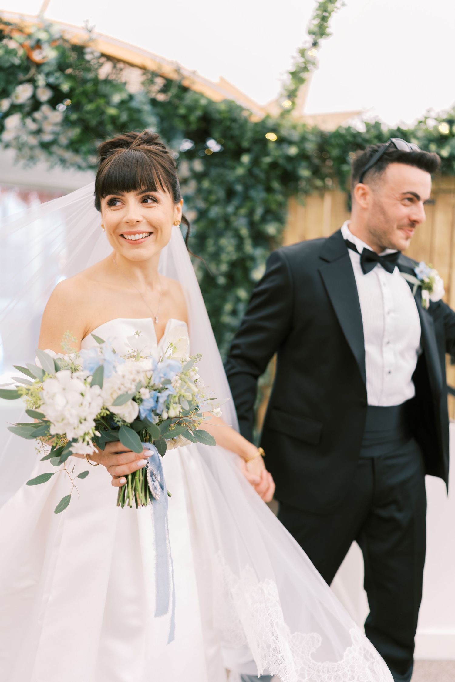 Smiling bride in strapless gown holds blue bouquet while groom in tuxedo walks beside her at outdoor wedding.
