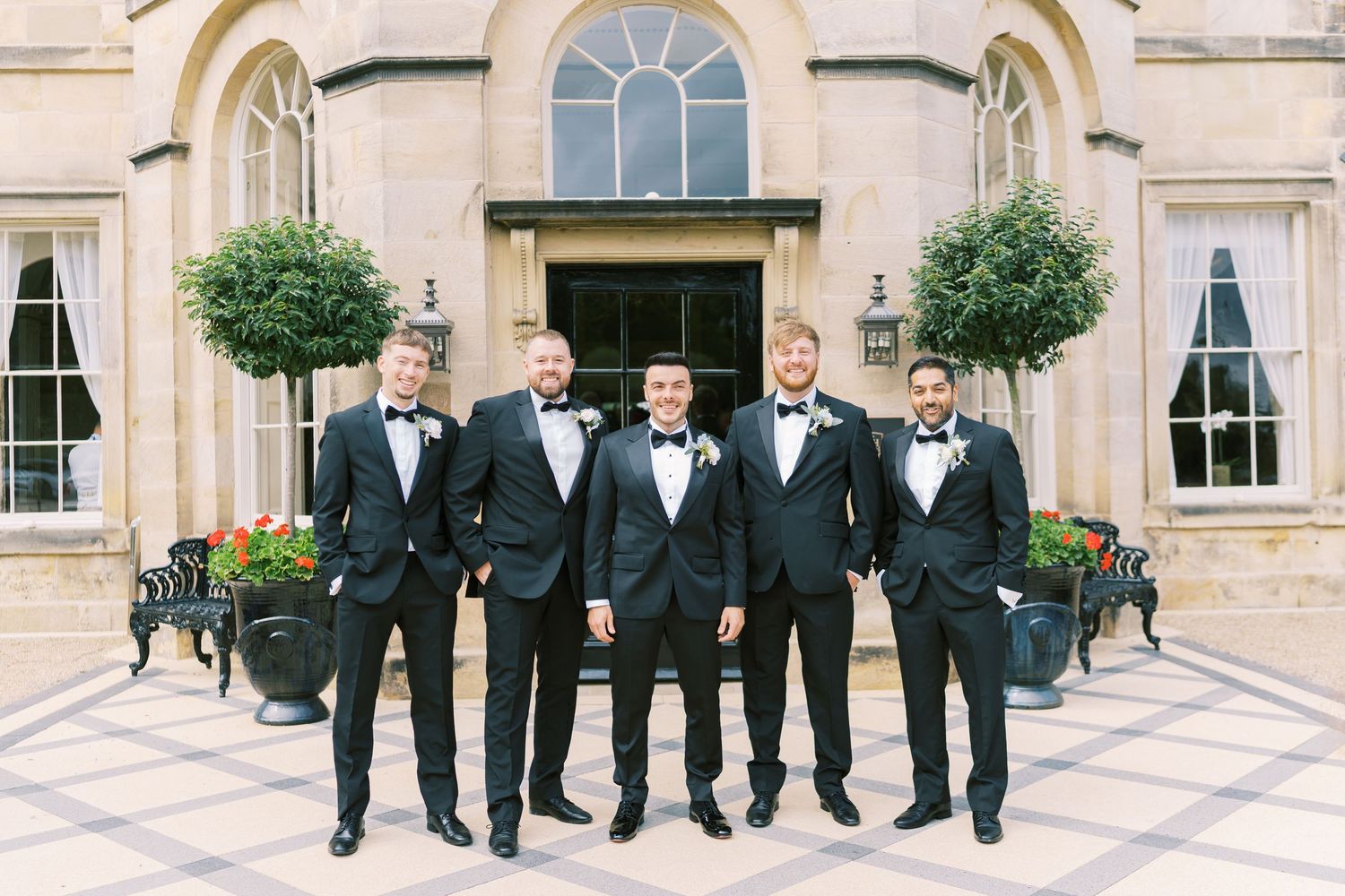 Five groomsmen in black tuxedos with bow ties pose outside an elegant stone building with topiary trees.