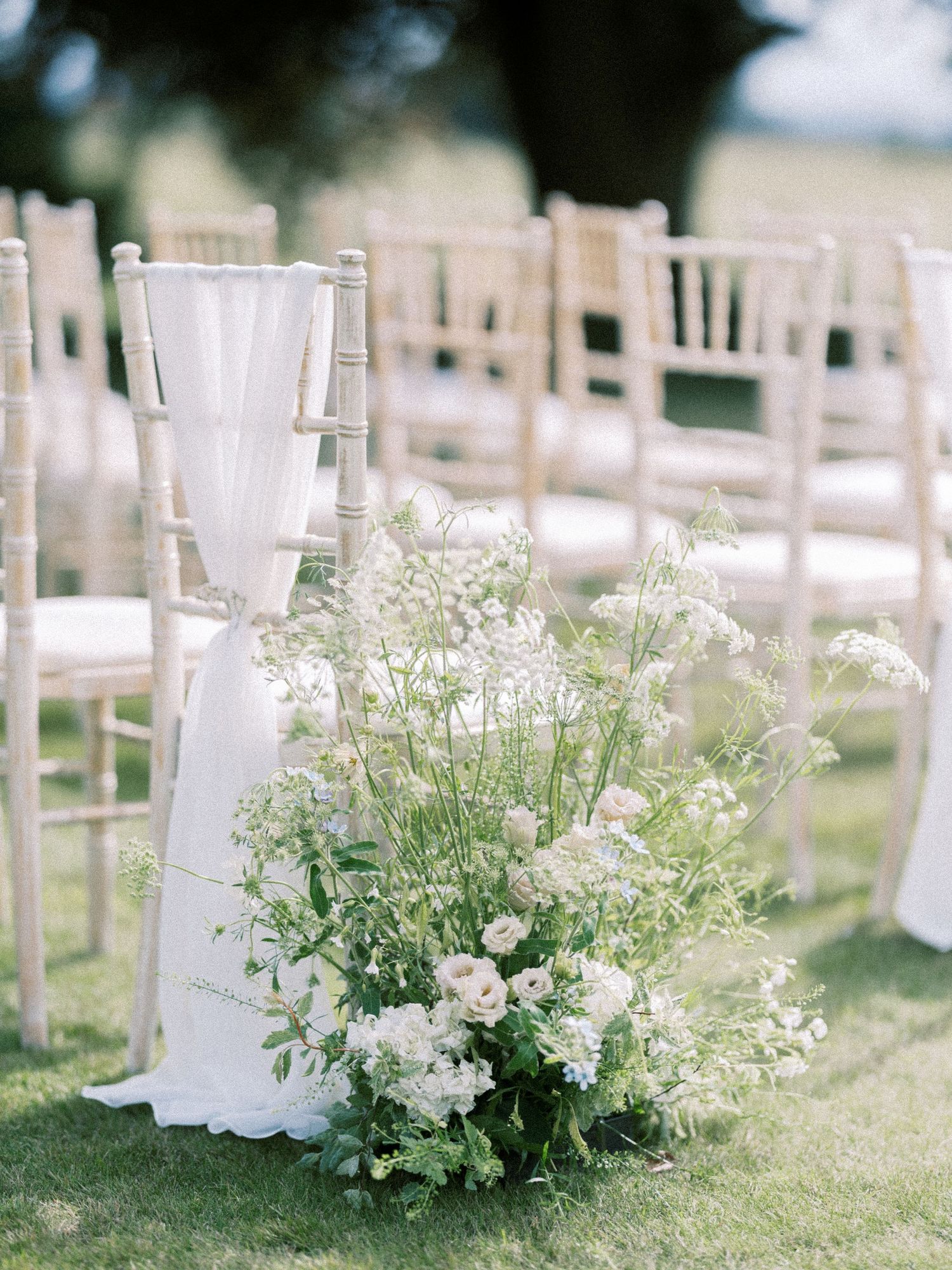 White floral aisle arrangement with wildflowers beside chiavari chairs at outdoor wedding ceremony.