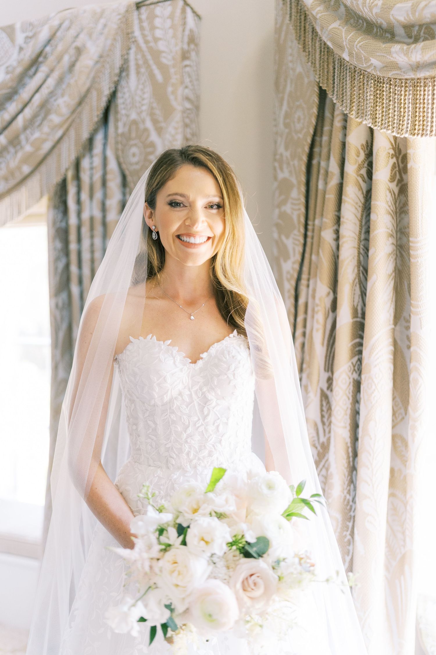 Smiling bride in strapless lace gown and veil holds white floral bouquet near elegant draped curtains.