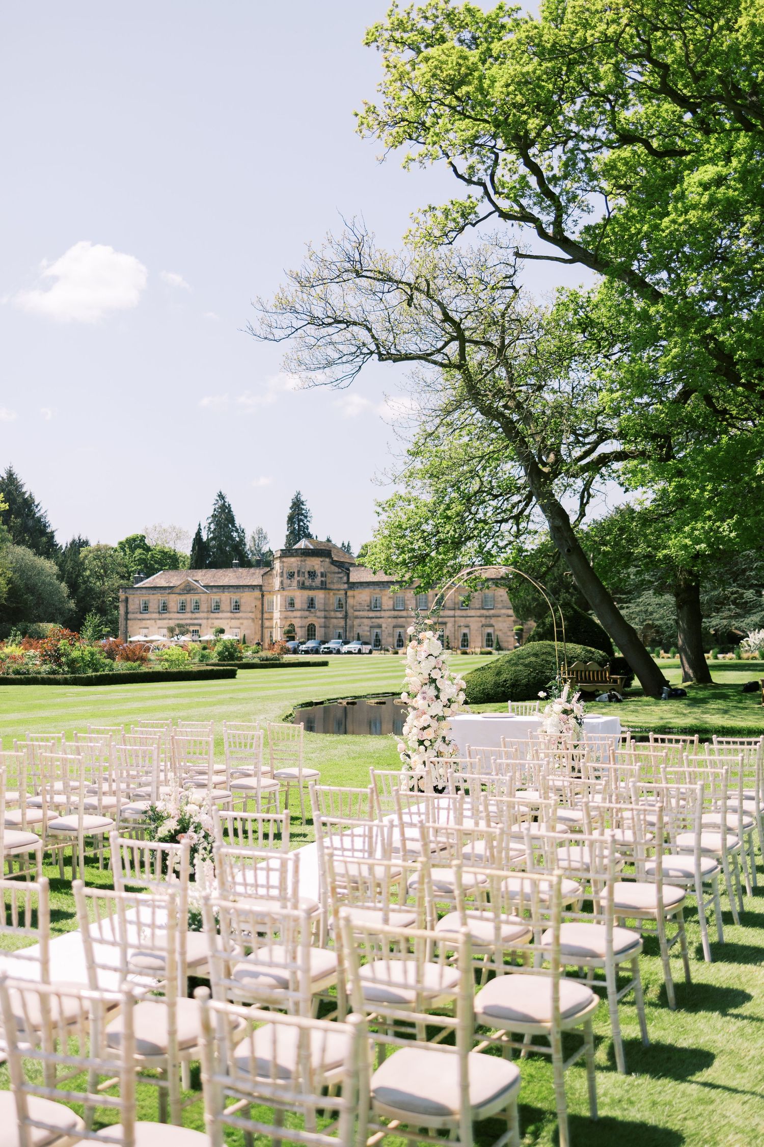 Elegant outdoor wedding ceremony setup with white Chiavari chairs on manicured lawn before a grand manor house.