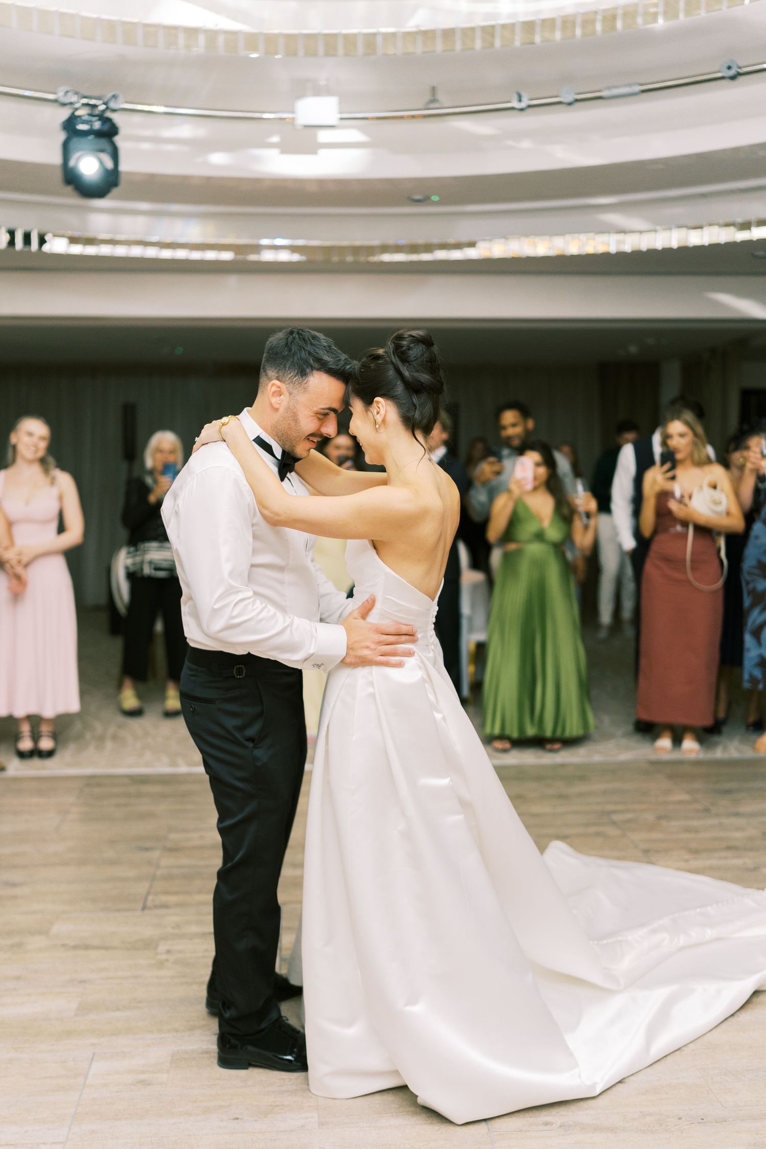 Bride and groom share their first dance at wedding reception, guests watching and photographing the romantic moment.