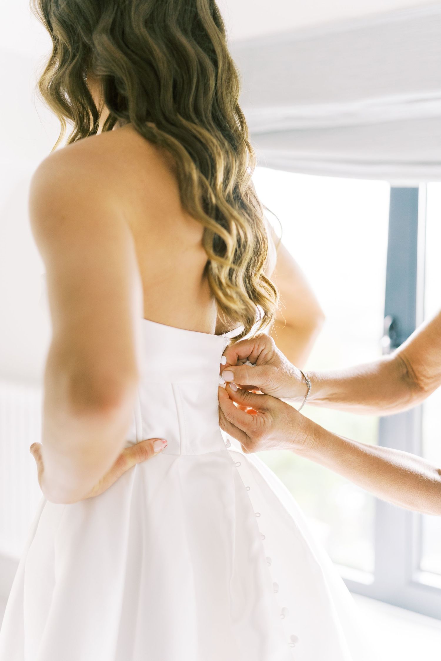 Bridesmaid fastening buttons on back of brides strapless white wedding gown before ceremony.