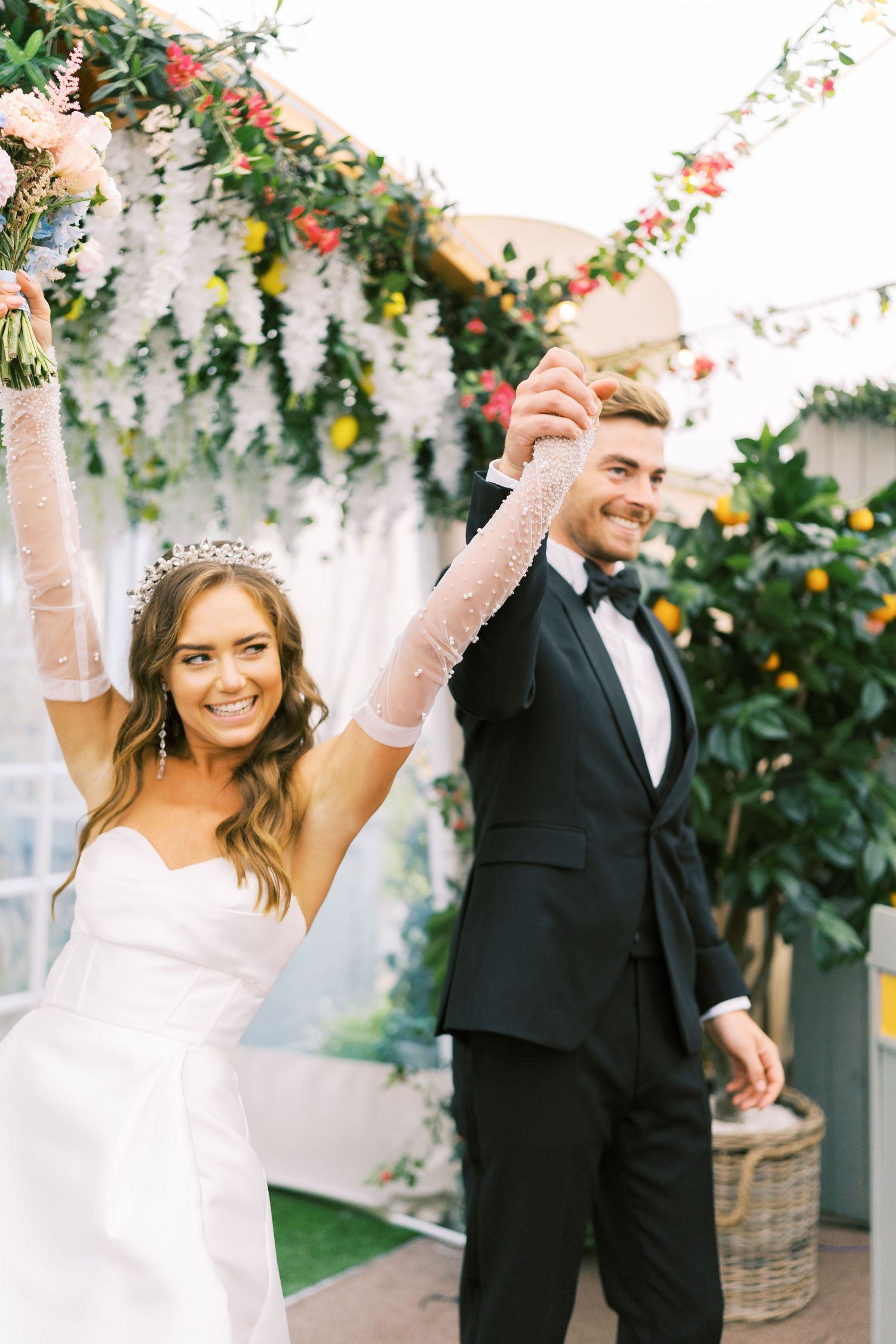 Joyful newlyweds celebrate under floral arch, bride in tiara and groom in black tuxedo raising hands.