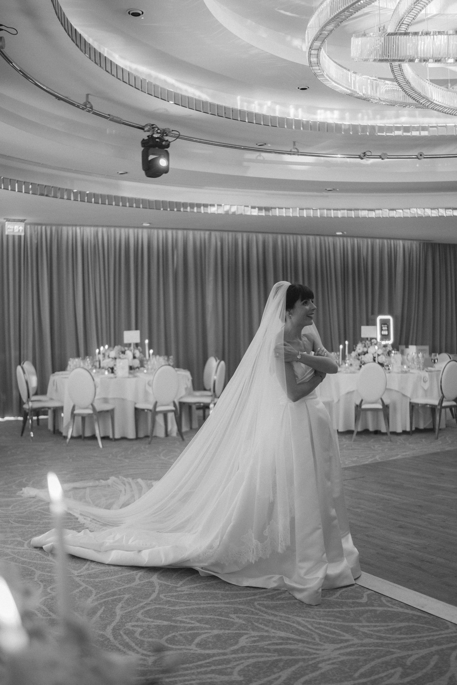 Black and white photo of a bride in a flowing gown and long veil standing in an elegant wedding reception ballroom.
