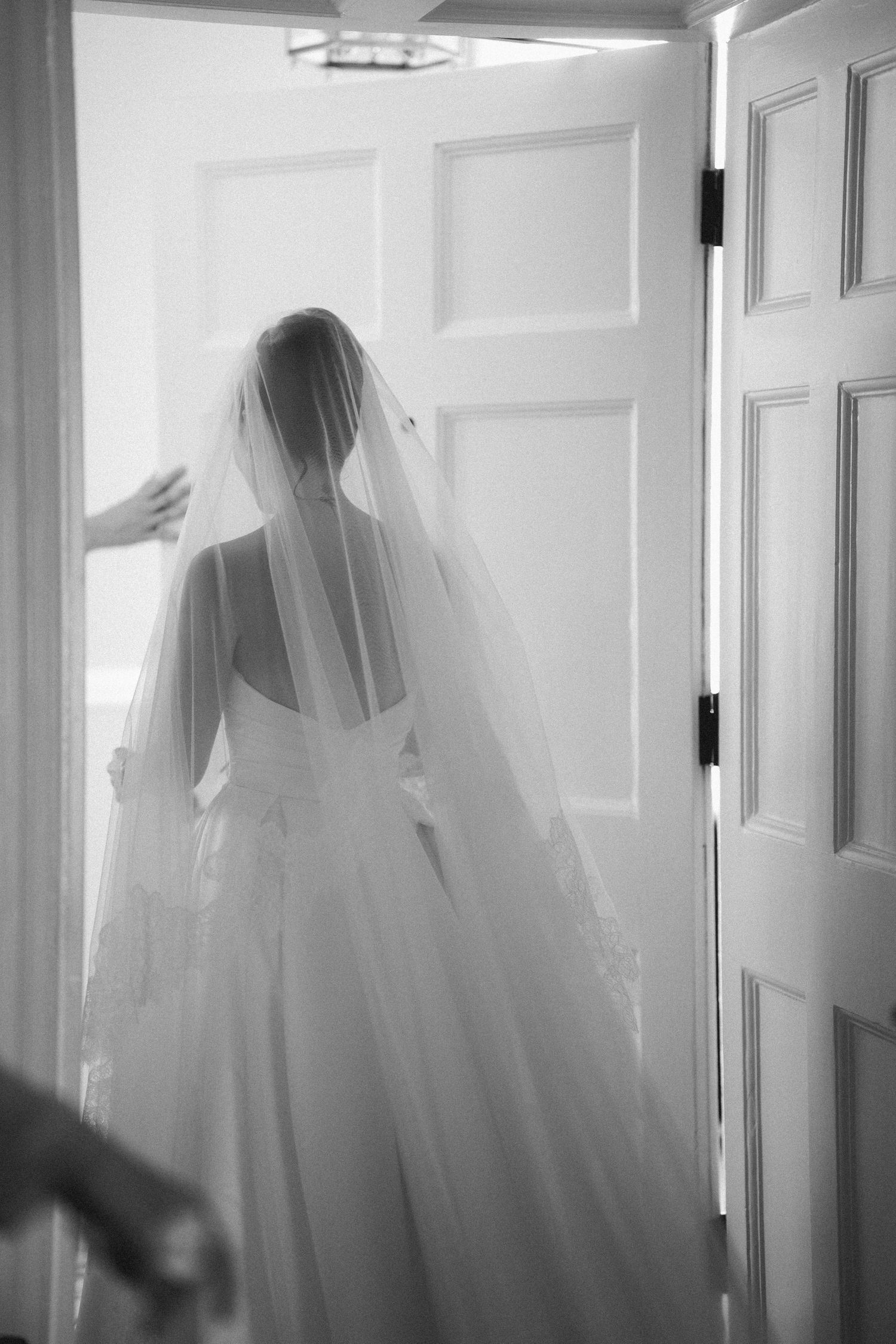 Bride in elegant wedding gown and long veil walking through doorway, black and white photo.