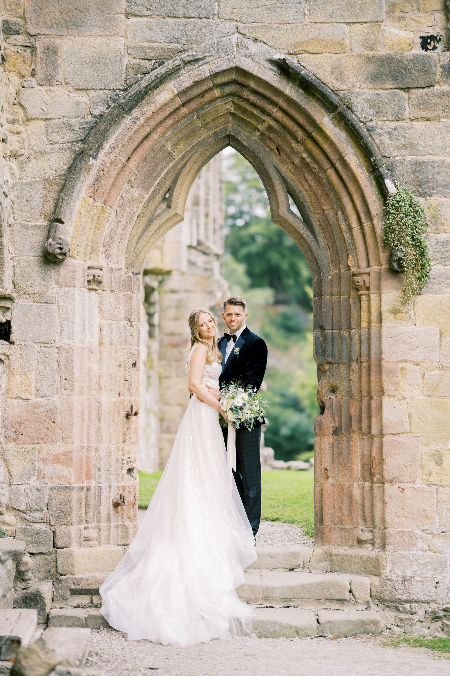 Bride and groom posing in a Gothic stone archway at historic ruins, bride in flowing white gown holding bouquet.