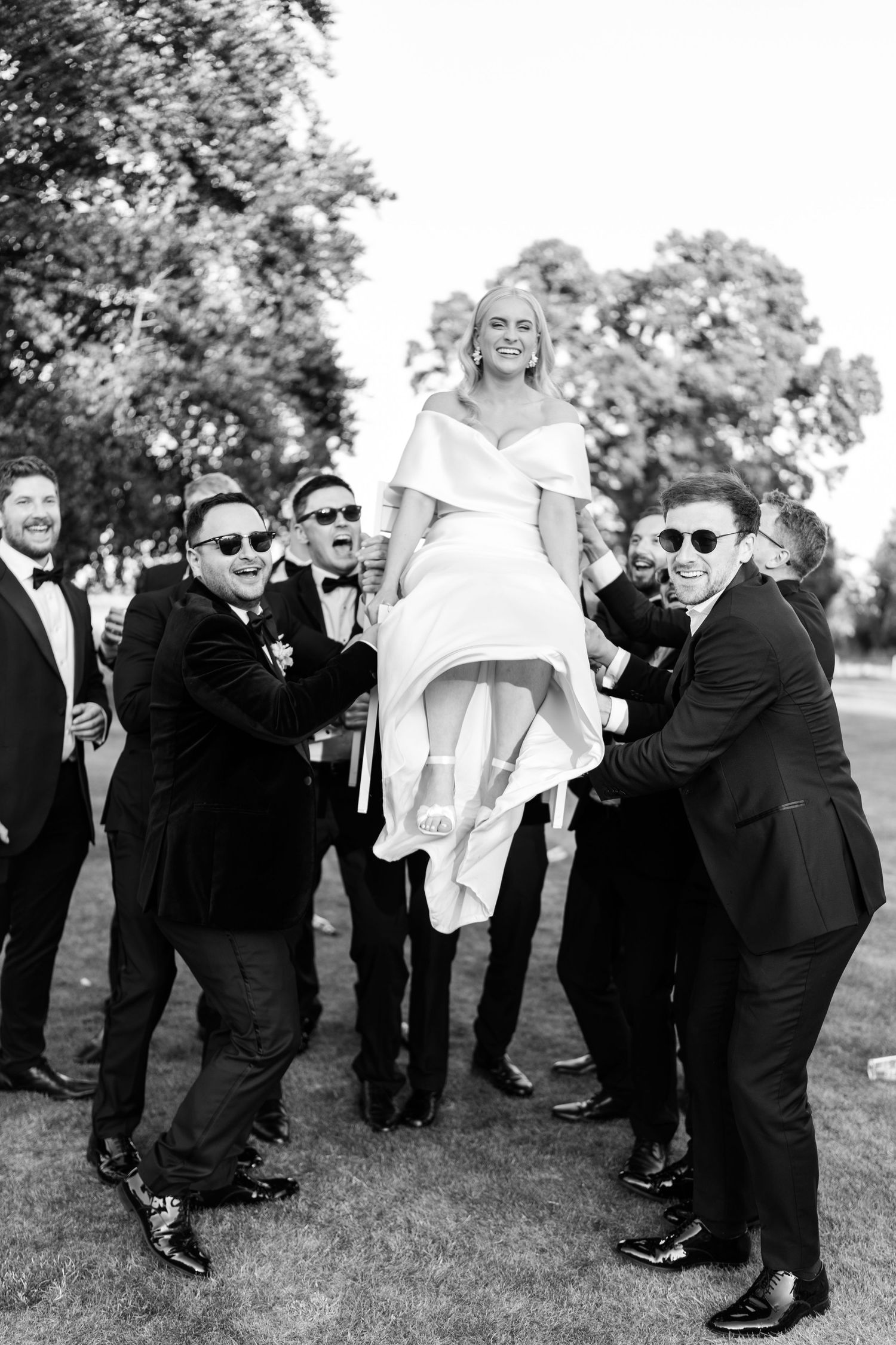 Black and white photo of groomsmen lifting the bride in her wedding gown outdoors.