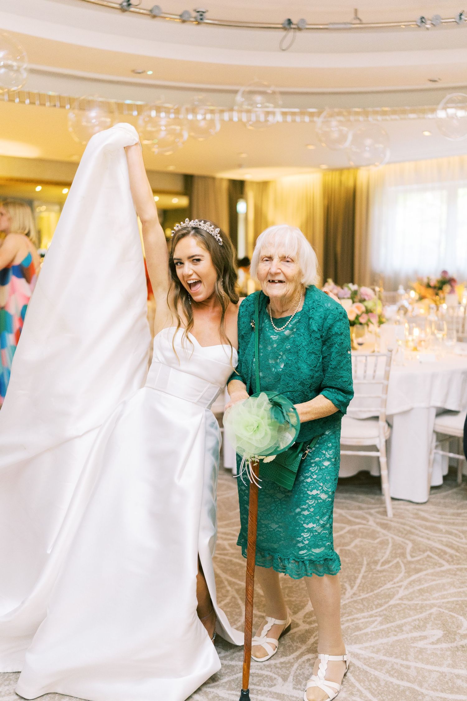 Bride in white gown and tiara laughs joyfully with elderly woman in teal lace dress at wedding reception.