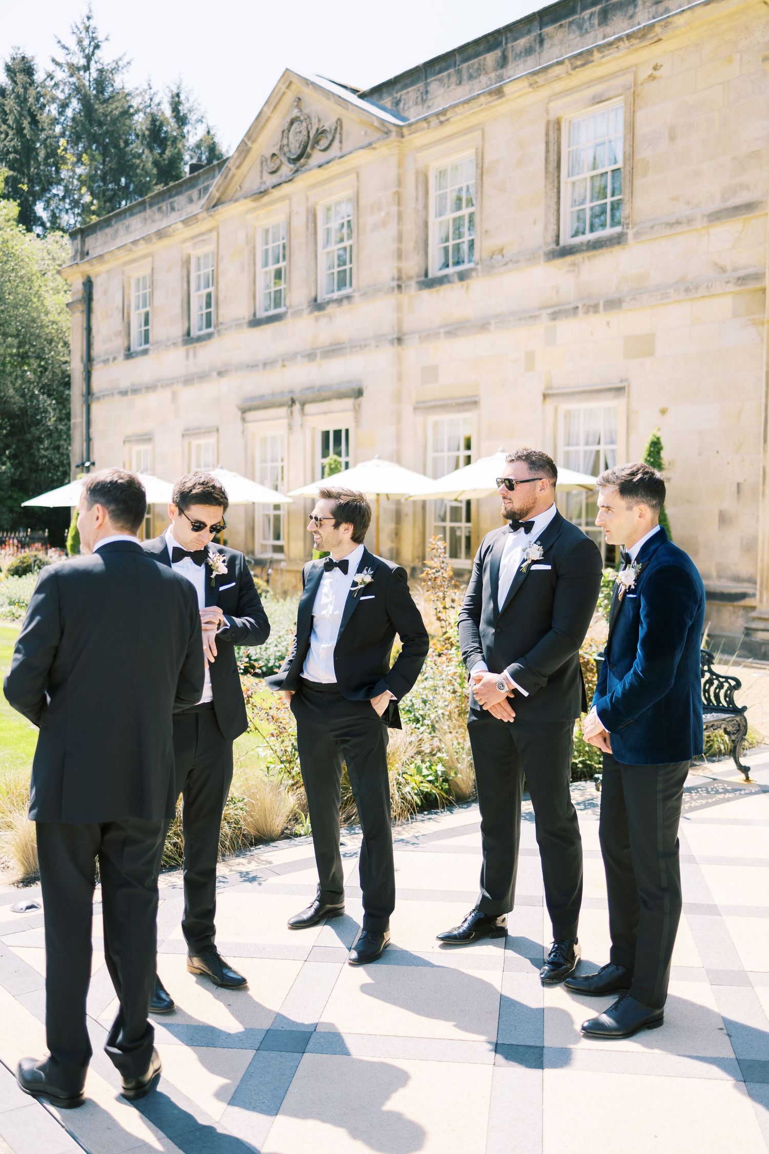 Groomsmen in black tuxedos with bow ties gather outside an elegant stone manor on a sunny wedding day.