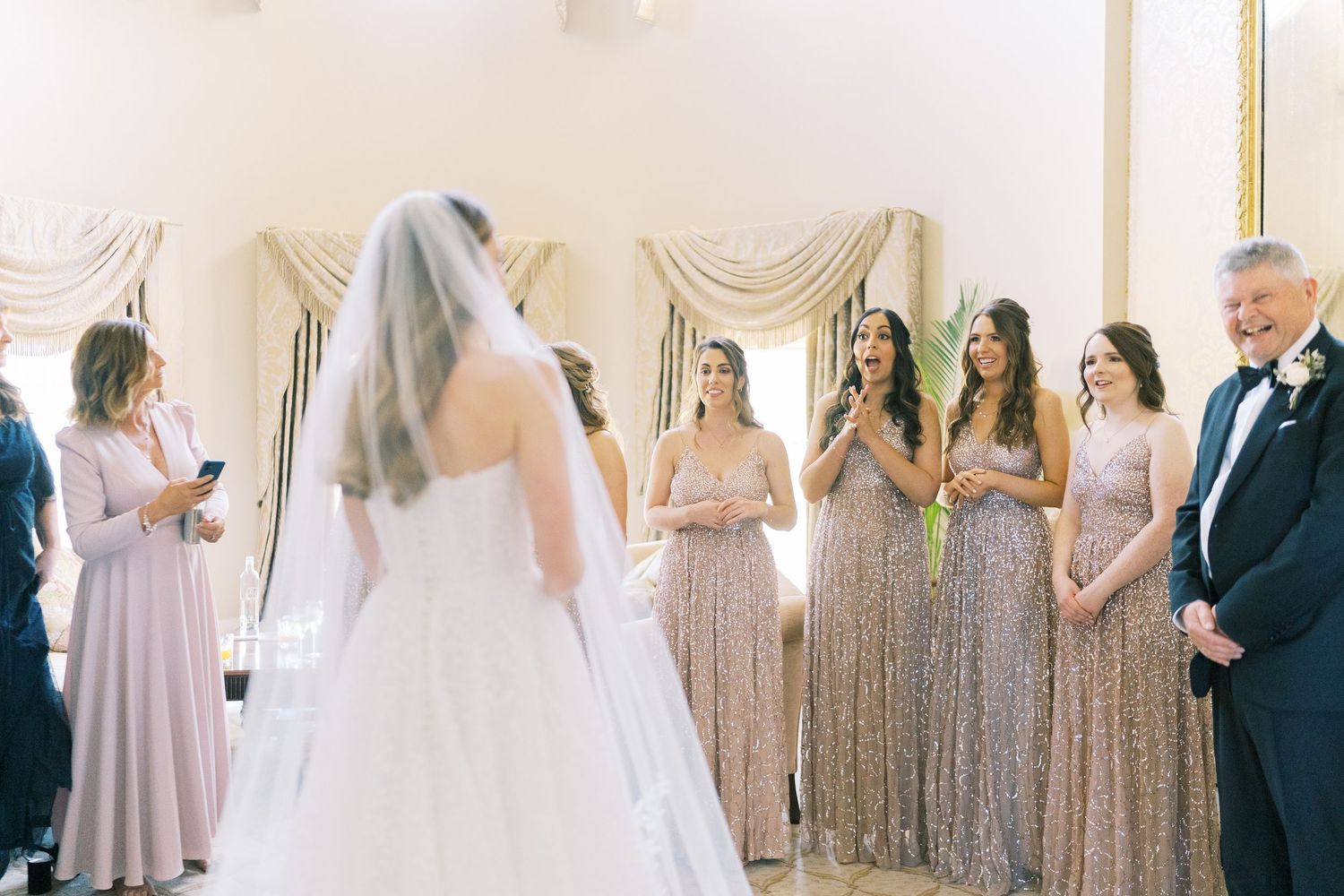 Bride in white gown and veil reveals look to bridesmaids in sparkly dresses and guests in elegant wedding venue.