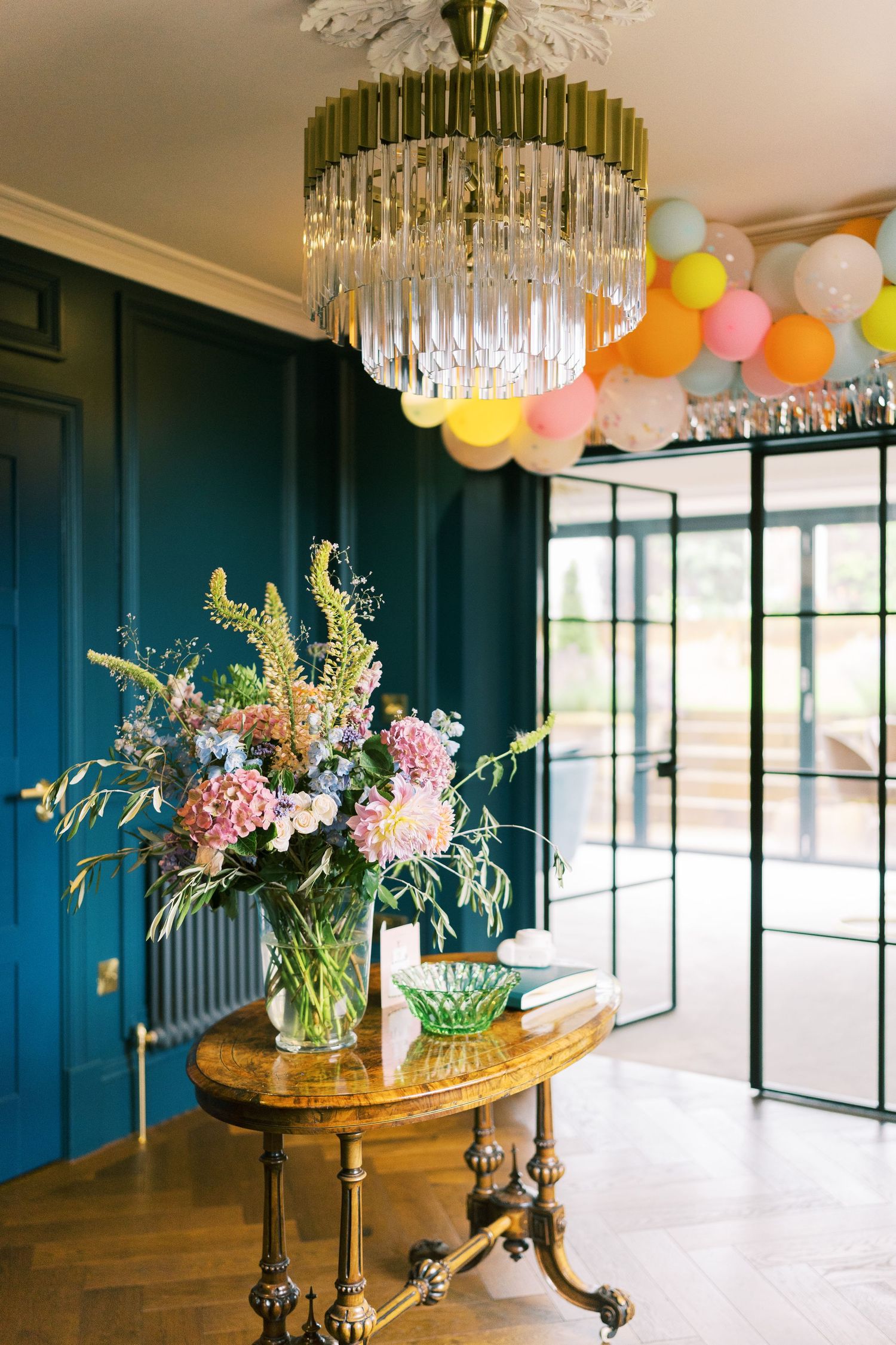 Elegant entryway with teal walls, crystal chandelier, colorful balloons, and a floral arrangement on a gold antique table.