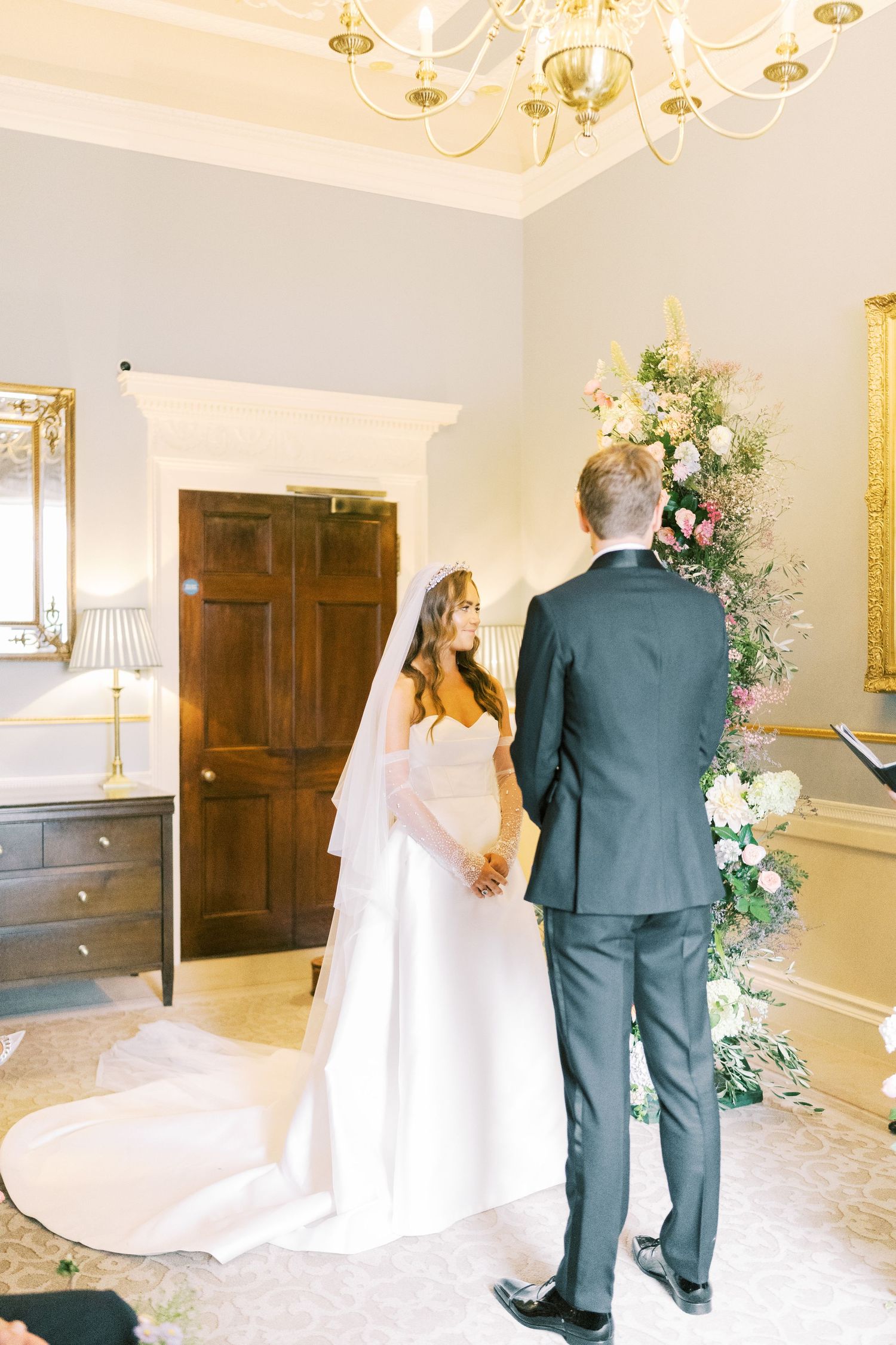 A bride and groom exchange vows at an elegant indoor wedding ceremony adorned with lush floral arrangements.