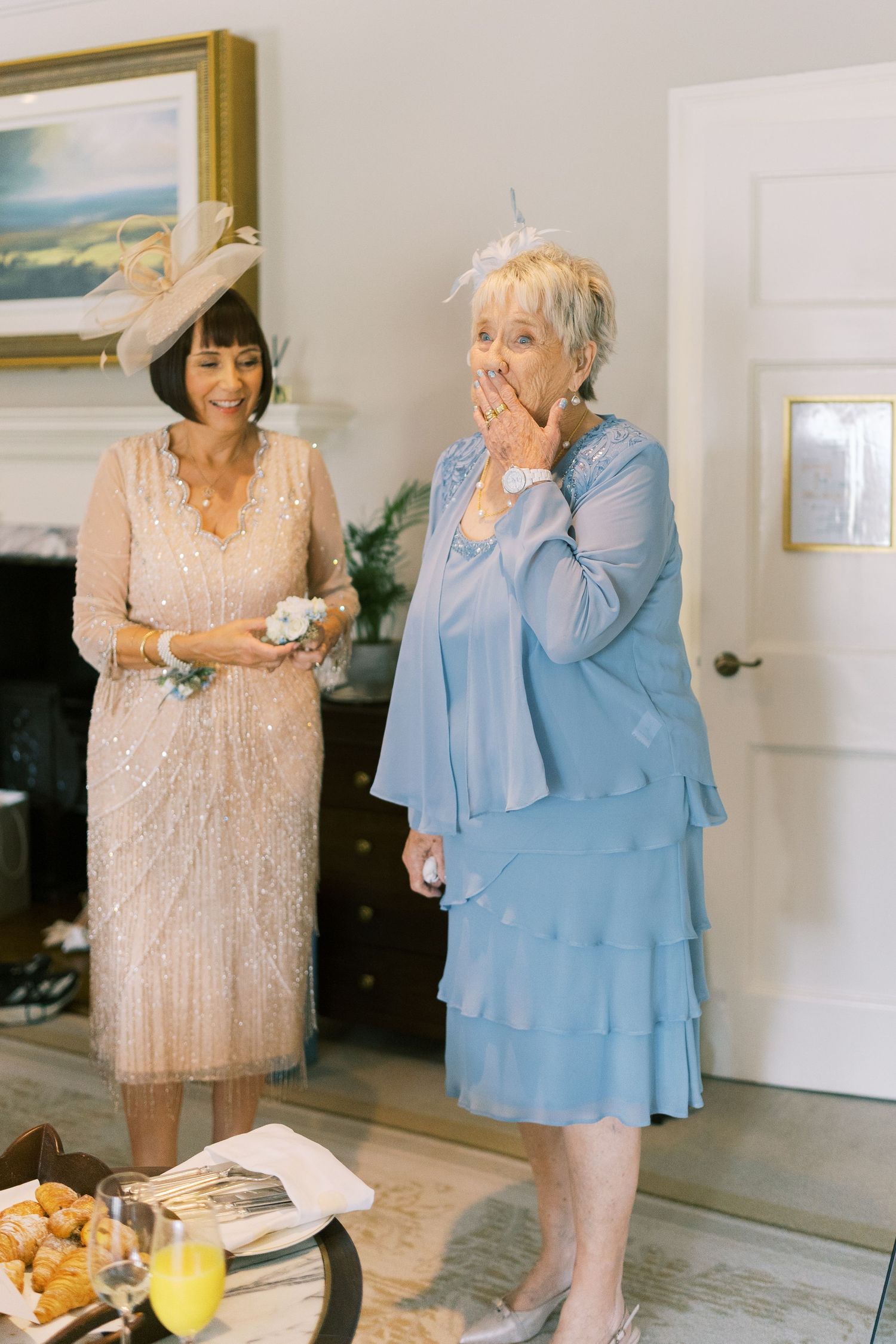 Two elegantly dressed women at a wedding, one in blush sequined dress, other in blue layered gown with surprised expression.