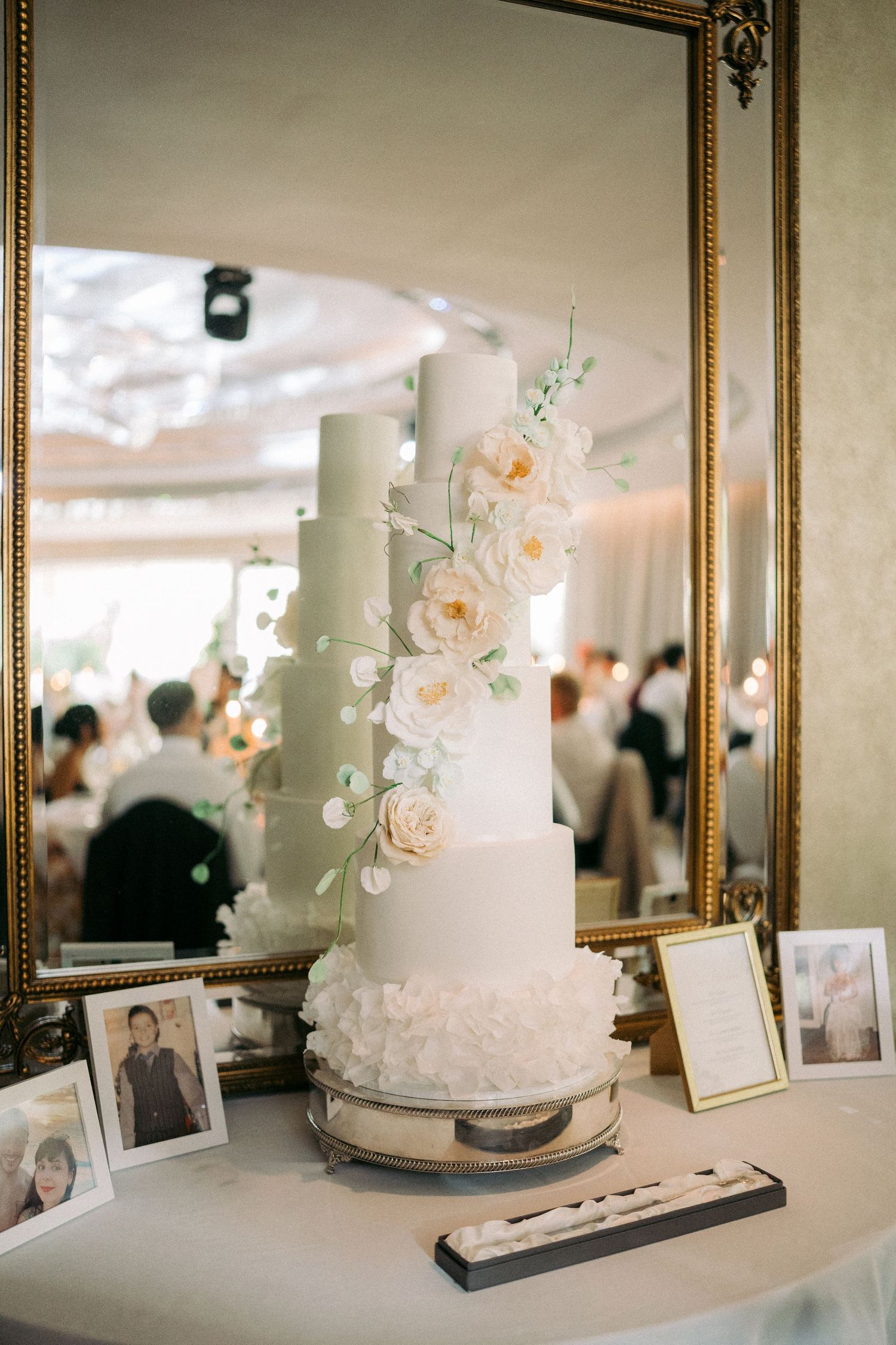 Elegant five-tier white wedding cake adorned with blush roses displayed on a table with framed photos at a reception.