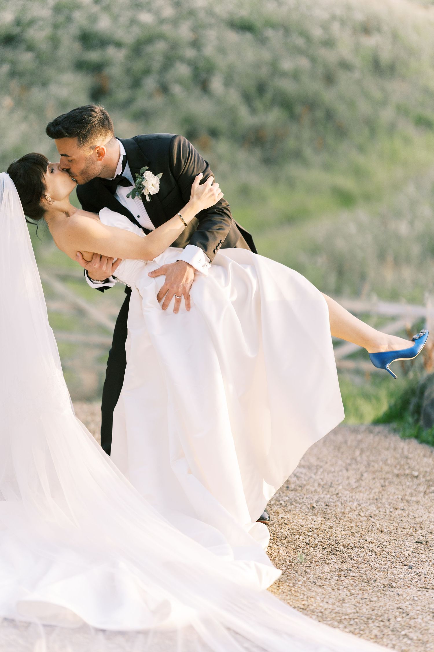 Groom in black tuxedo dipping bride in white wedding dress and blue heels for a romantic kiss outdoors.