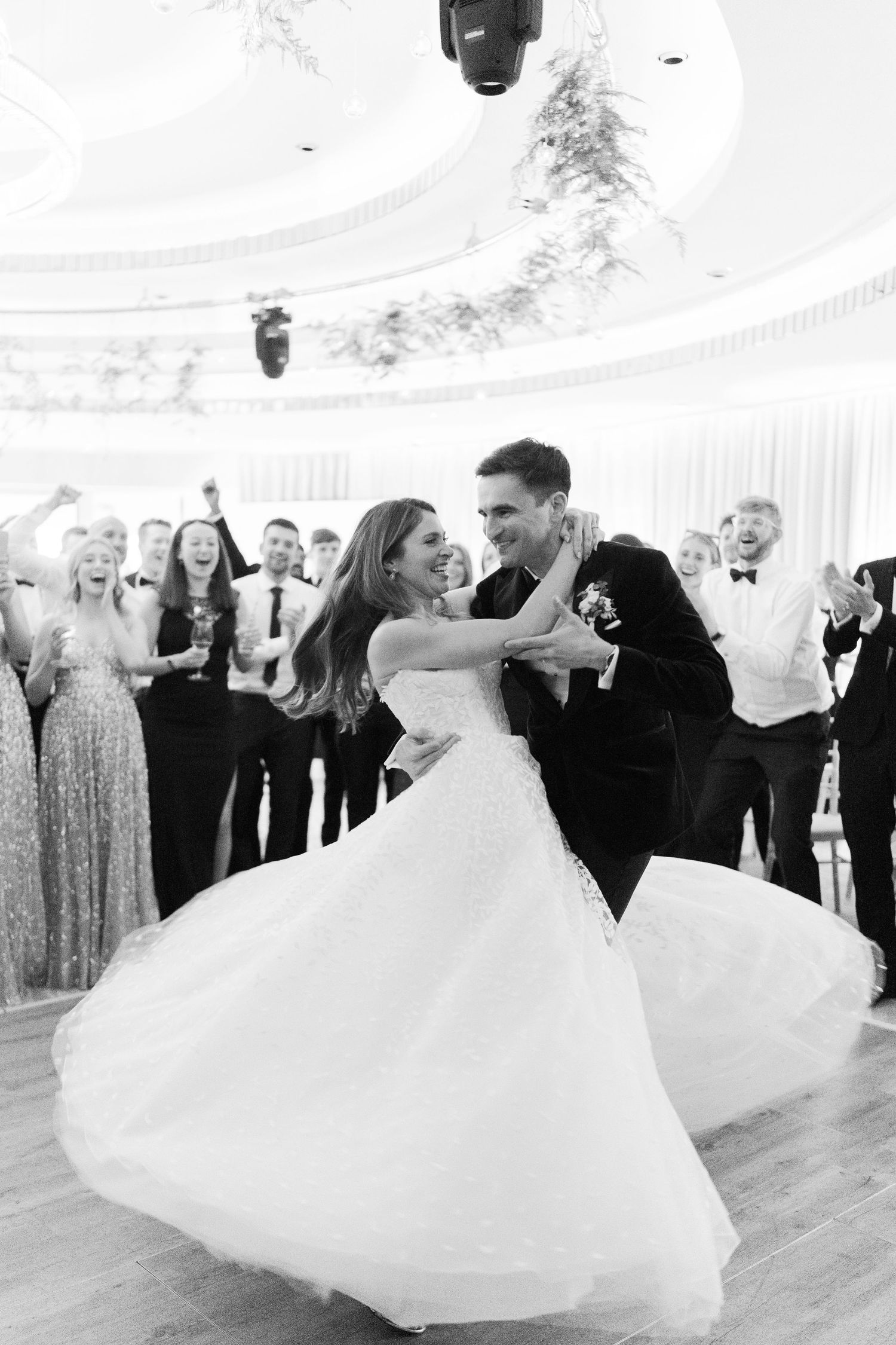Bride and groom share first dance at elegant wedding reception while guests celebrate in black and white photo.