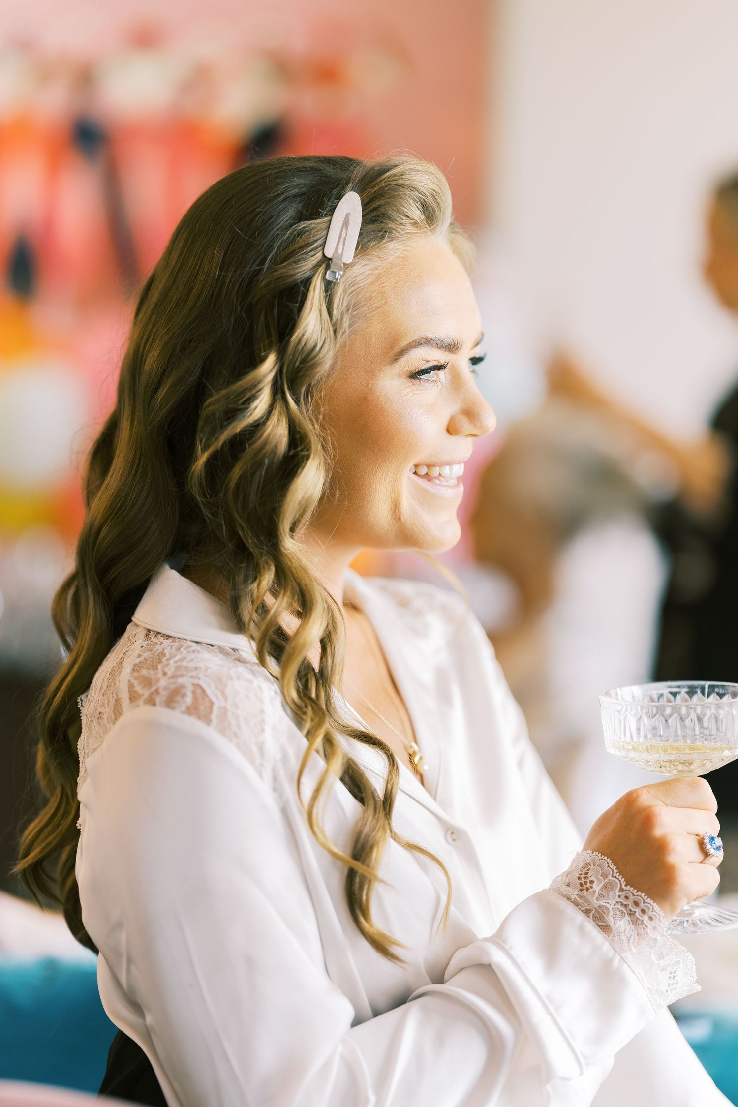 Smiling bride in white lace robe holding crystal champagne glass with elegant wavy hair.