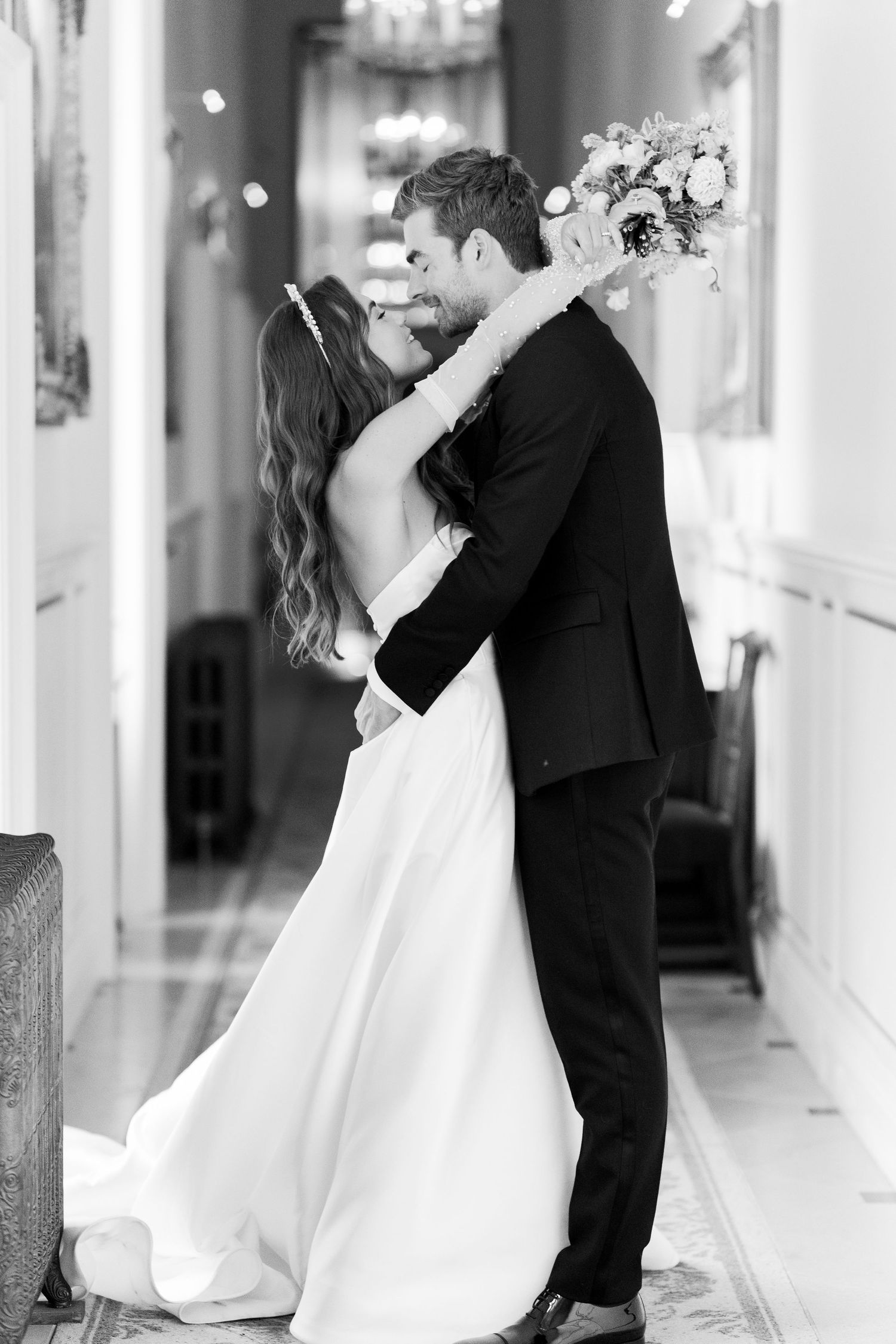 Black and white photo of bride and groom embracing in elegant hallway, bride holding bouquet.