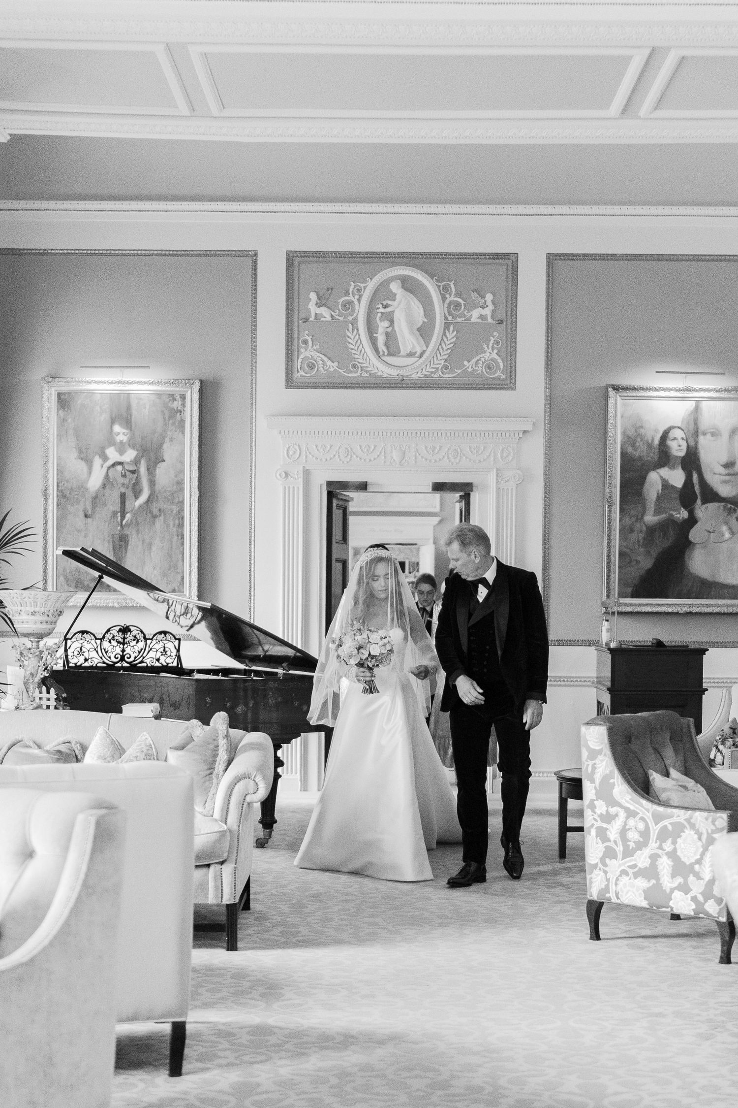 Black and white photo of a bride walking with her father through an elegant room before her wedding ceremony.