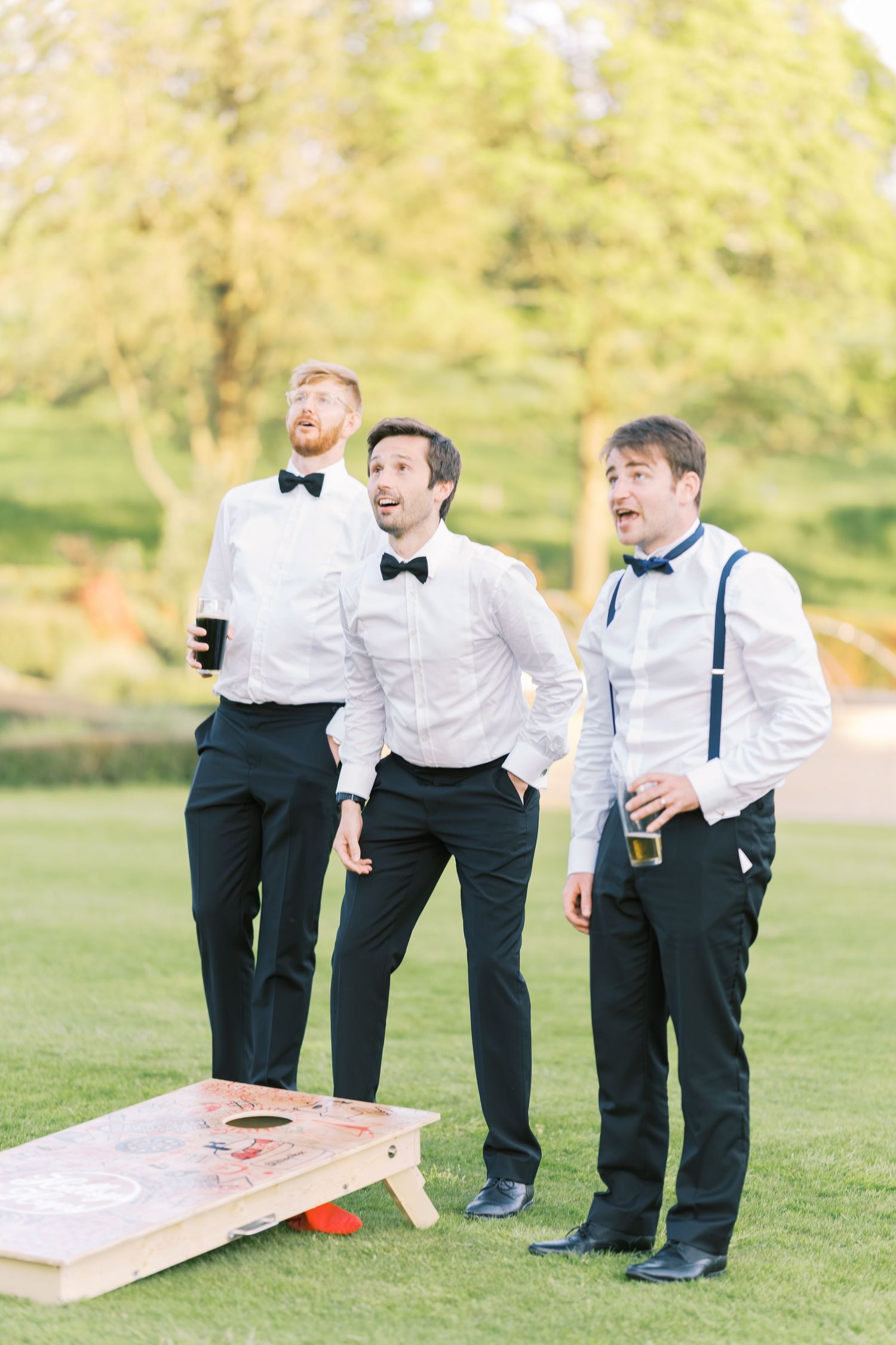Three groomsmen in bow ties and suspenders playing cornhole lawn game at an outdoor wedding reception.