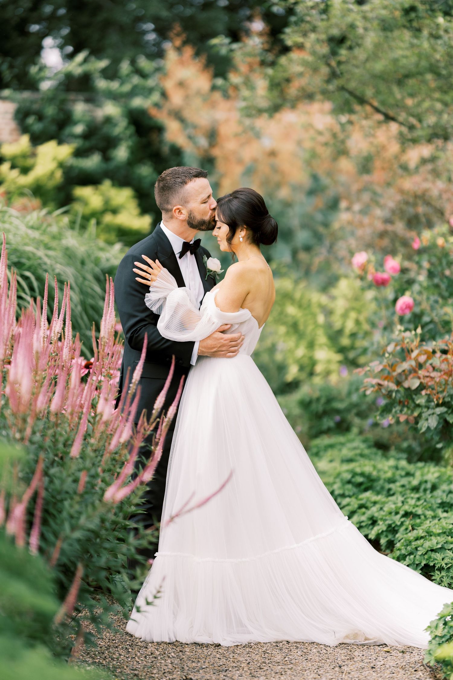 Bride and groom share a romantic moment in a lush garden, bride in flowing white off-shoulder gown.