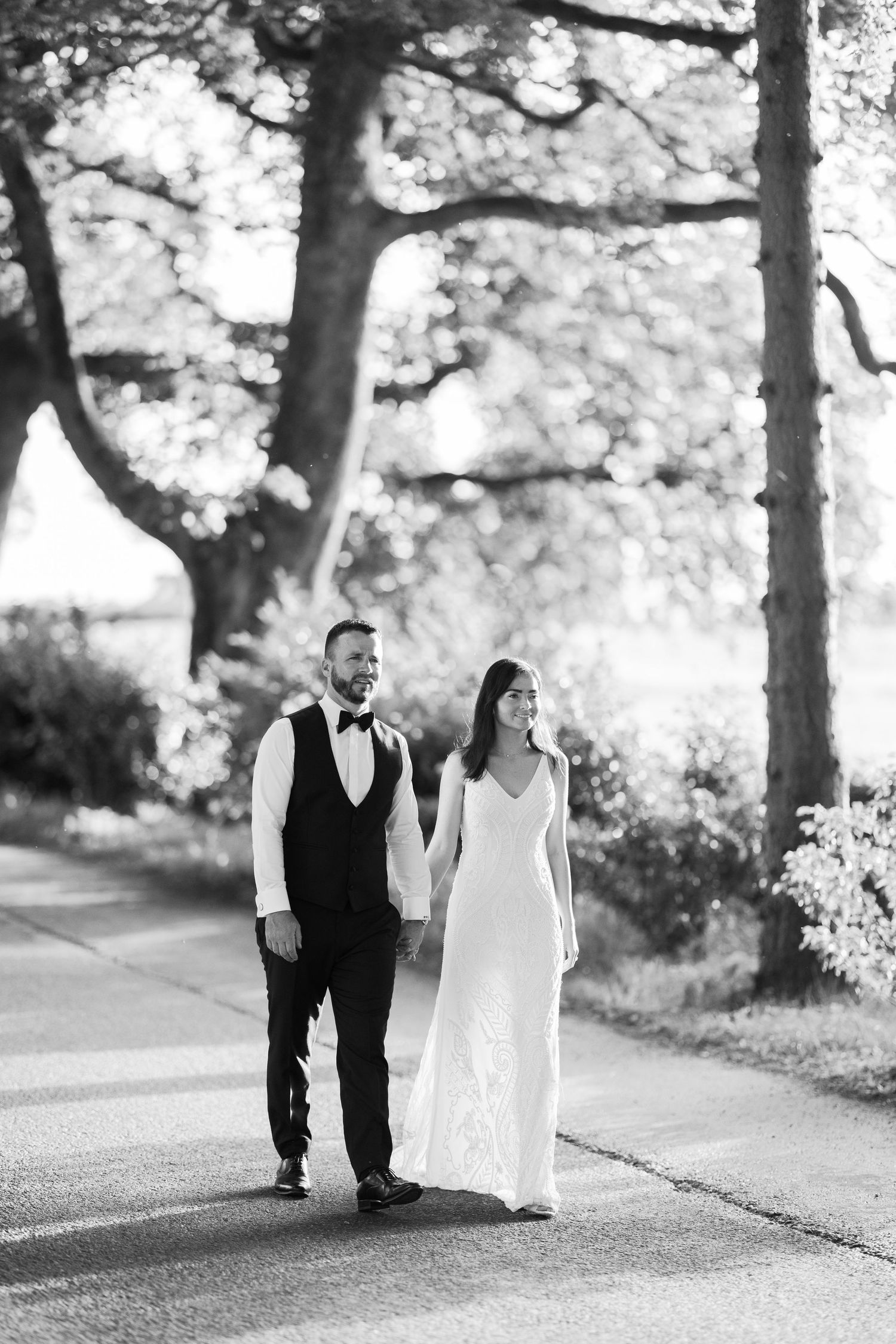Black and white photo of a bride and groom walking hand in hand along a tree-lined path on their wedding day.