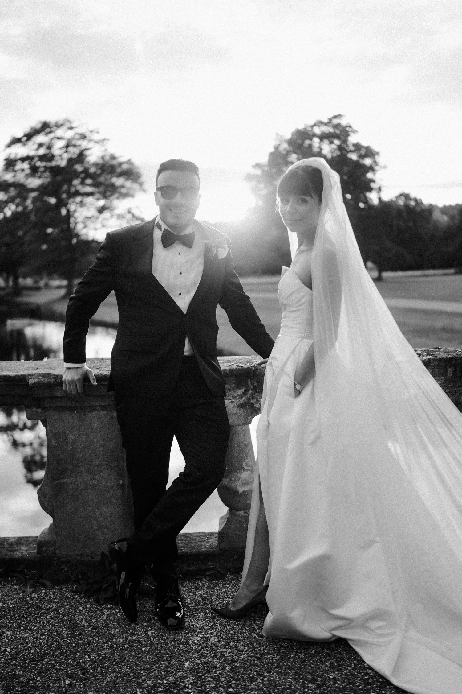 Black and white photo of bride in veil and groom in tuxedo posing by stone balustrade at sunset.