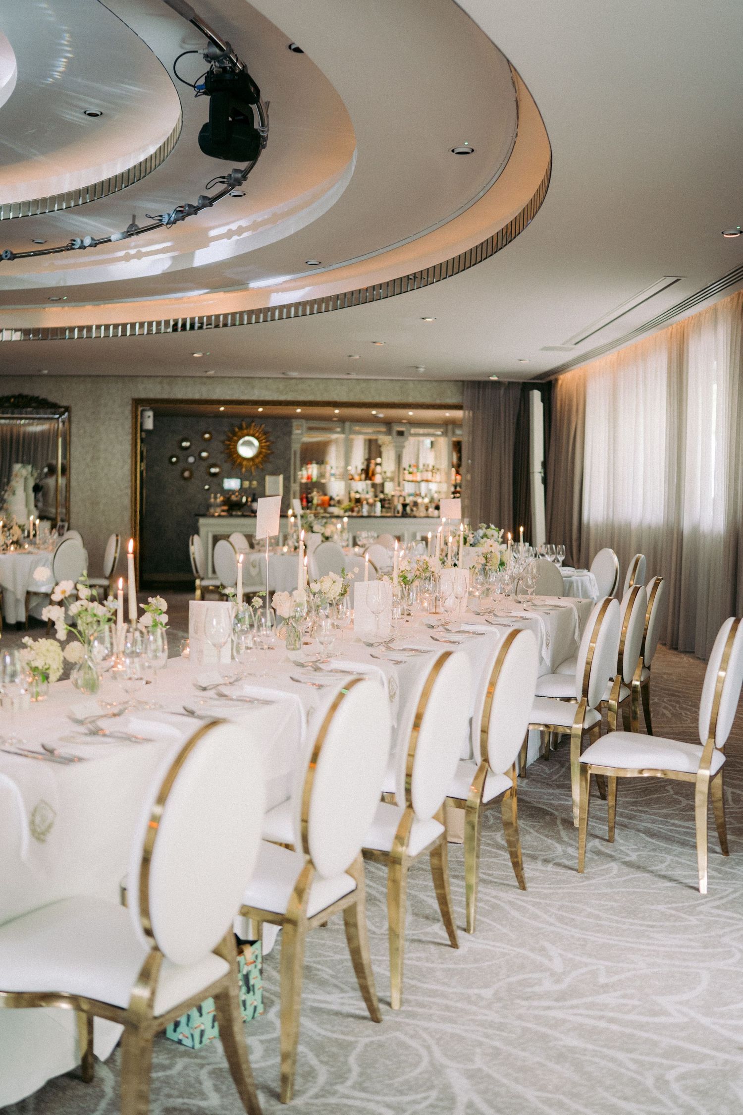 Elegant wedding reception hall with white and gold chairs, long dining tables, floral centerpieces, and a circular ceiling detail.