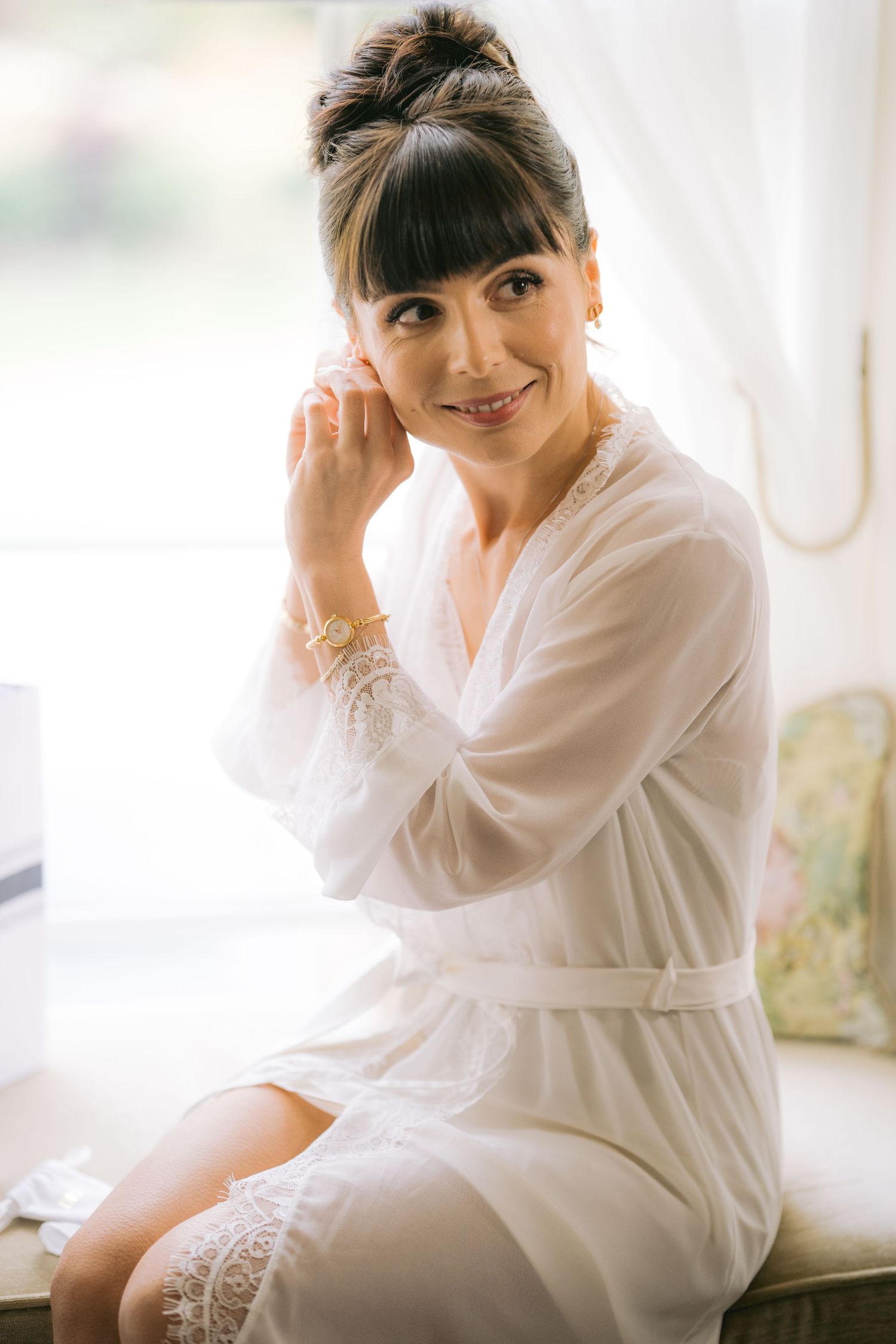 Bride in white lace robe smiling while putting on earrings before her wedding.