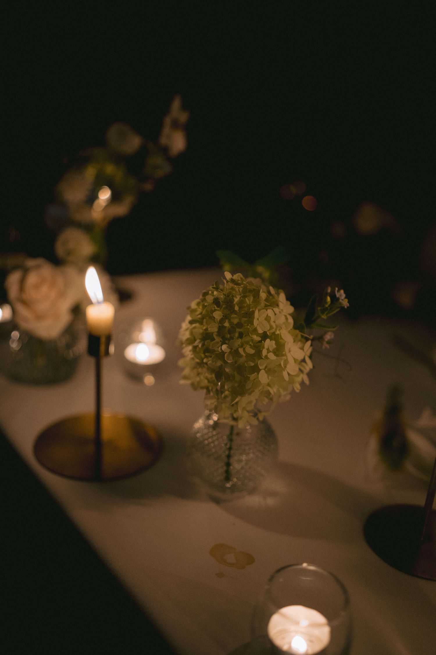 Romantic candlelit wedding table with white hydrangea in glass vase and glowing tea lights in dark setting.