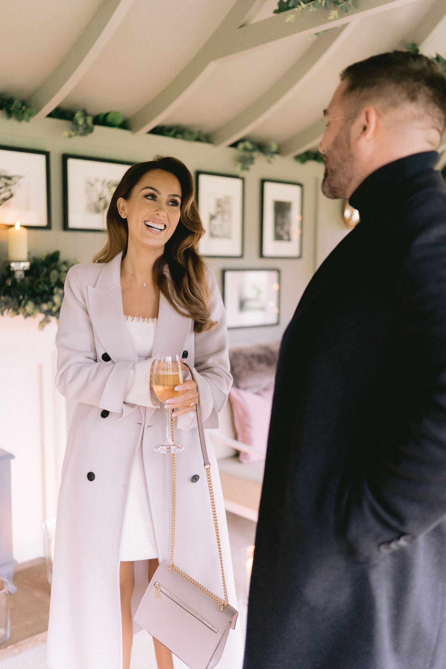 Stylish woman in a beige coat holding a wine glass and laughing in a charming decorated holiday interior.