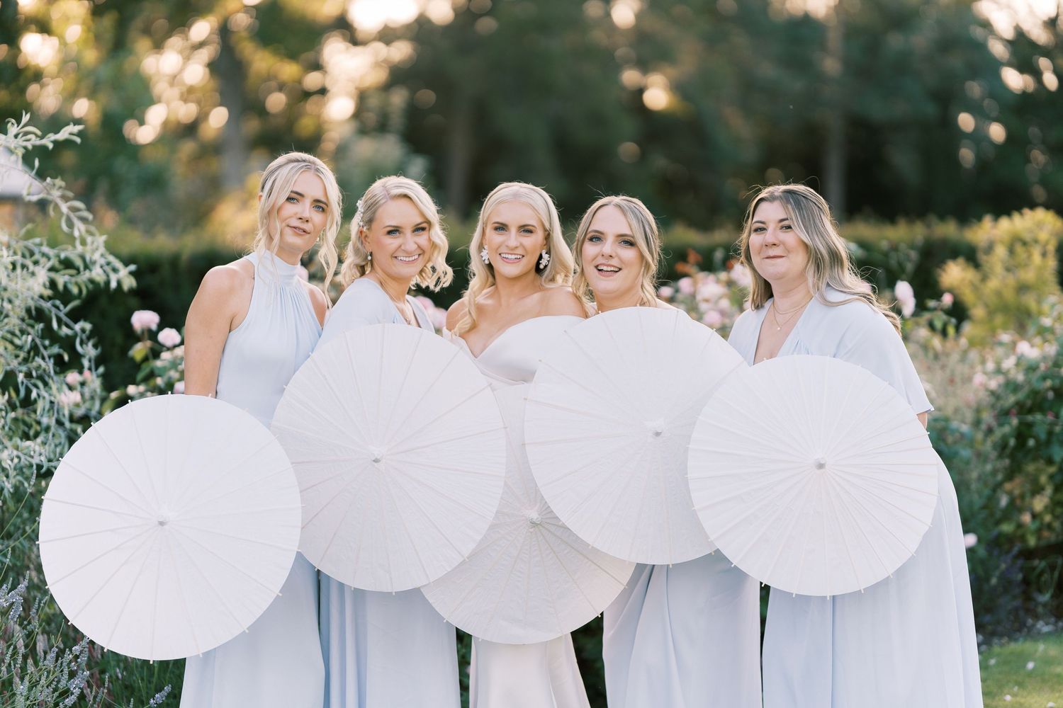 Bride and four bridesmaids in light blue dresses holding white parasols in a garden setting.